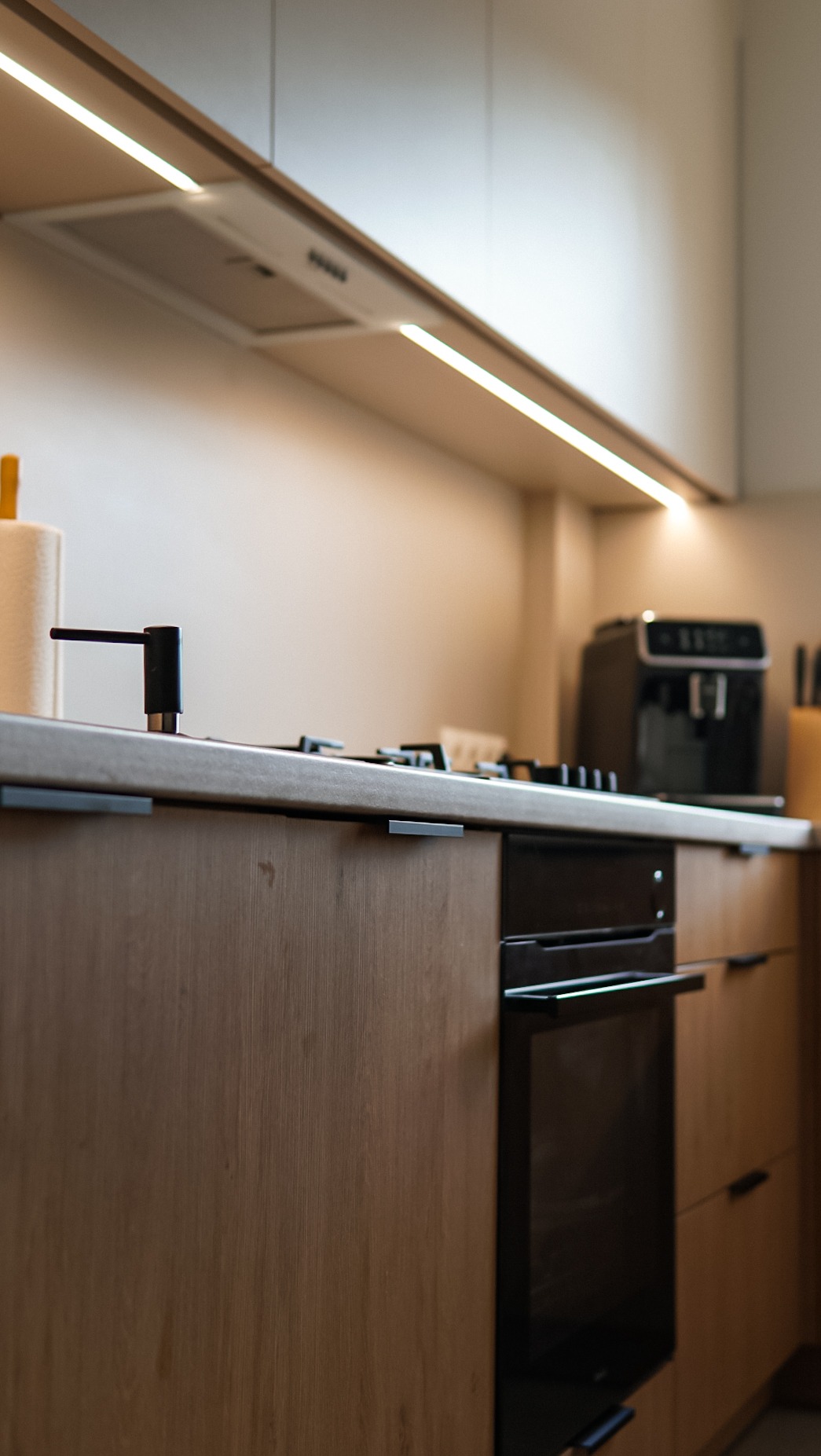 Nowoczesna kuchnia: zabudowa z drewnianymi frontami, czarny piekarnik i oświetlenie LED pod szafkami. Minimalistyczny design z ekspres do kawy w tle.