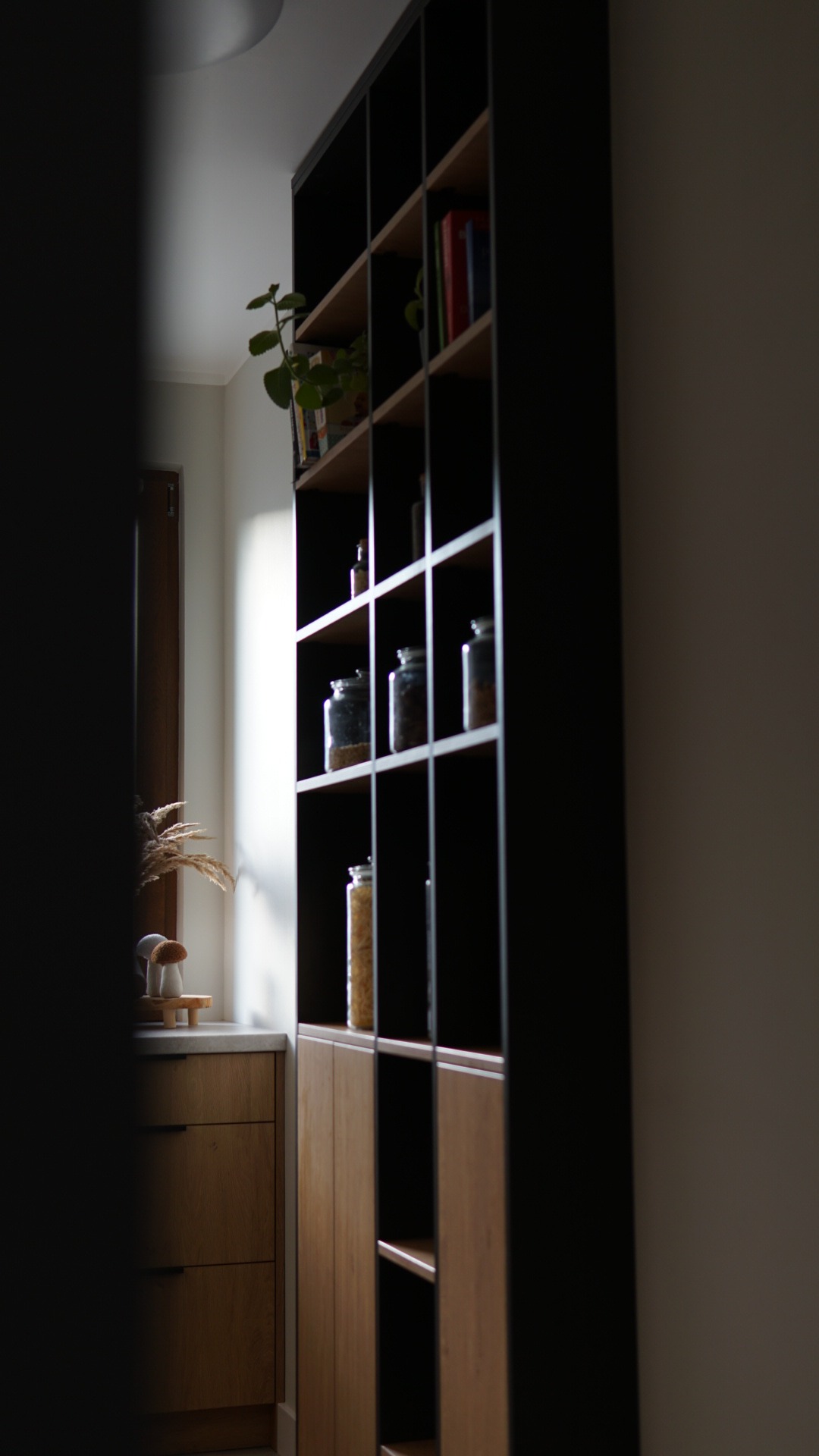 Częściowy widok na zabudowę kuchenną z ciemną, regałową szafką na przyprawy i drewnianymi frontami szafek dolnych, z boku okno z dekoracjami.