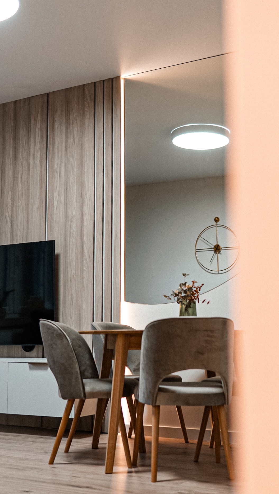 Nowoczesna jadalnia z drewnianym stołem i szarymi krzesłami. Na ścianie lustro z podświetleniem LED. Minimalistyczny zegar i kompozycja kwiatowa dodają elegancji.