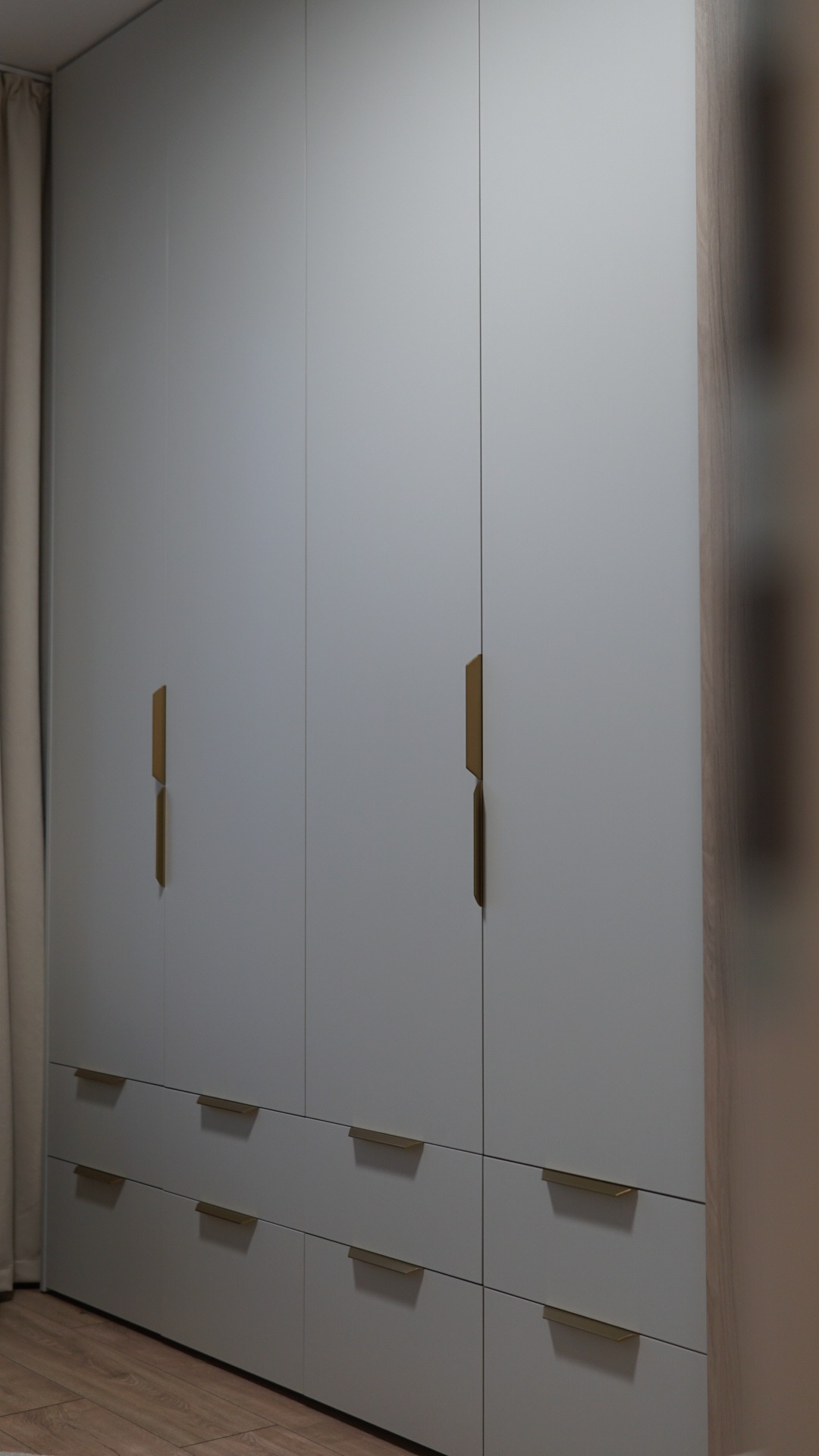 Elegancka, szara szafa wnękowa z złotymi, minimalistycznymi uchwytami, podzielona na część z drzwiami i dolne szuflady. Widoczne drewniane wykończenie boku szafy.