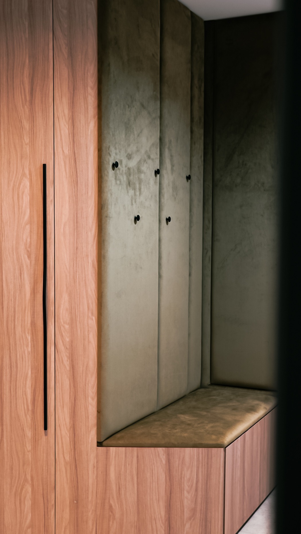 Drewniana szafa z długim, czarnym uchwytem sąsiaduje z tapicerowaną wnęką z siedziskiem i wieszakami. Nowoczesny design, stonowana kolorystyka.