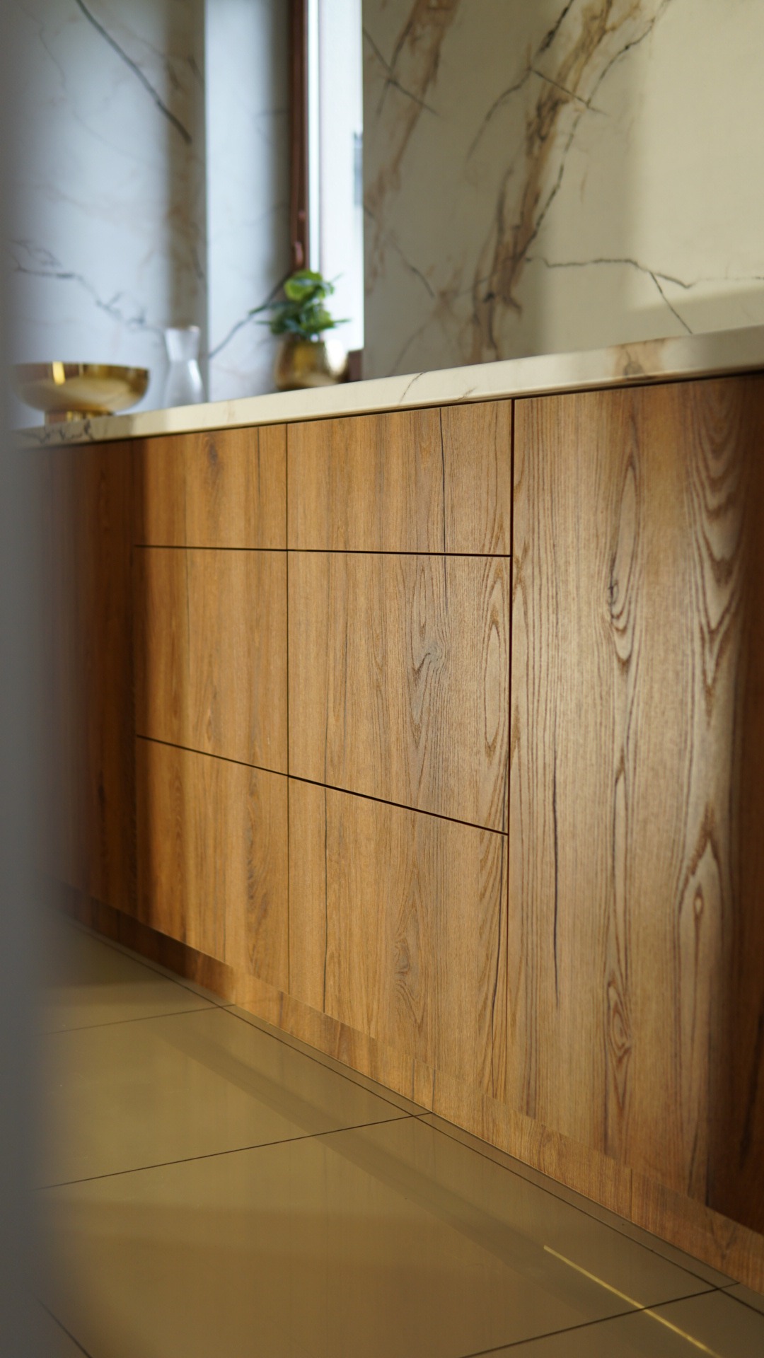Fronty szafek kuchennych z drewna o widocznym usłojeniu, jasny blat i marmurowe wykończenie ściany. Minimalistyczny styl, ciepłe oświetlenie.