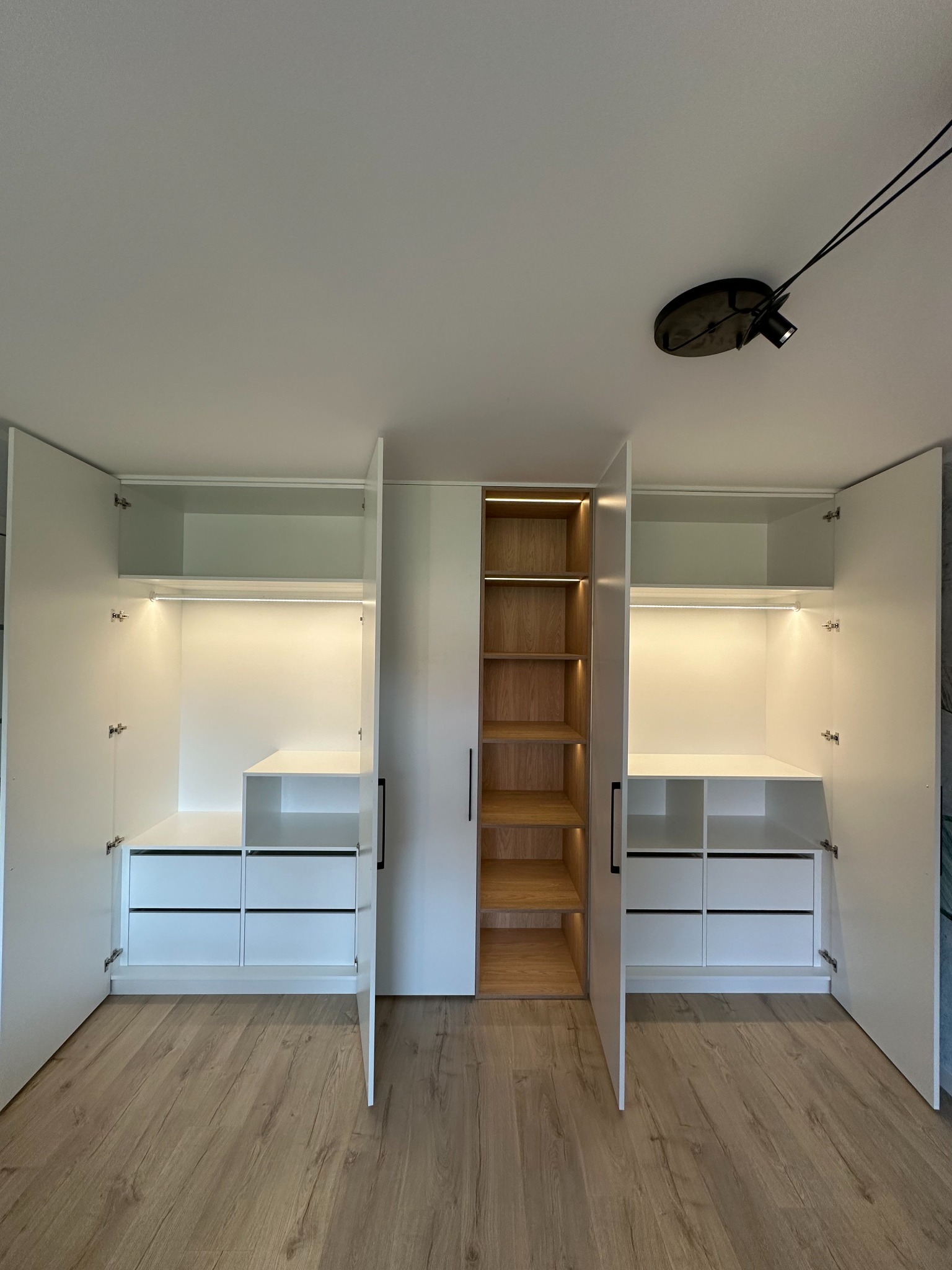 Nowoczesna szafa wnękowa z białymi frontami, oświetleniem LED i drewnianymi półkami. Otwarta przestrzeń z szufladami i półkami. Widoczne detale wykończenia i oświetlenie.