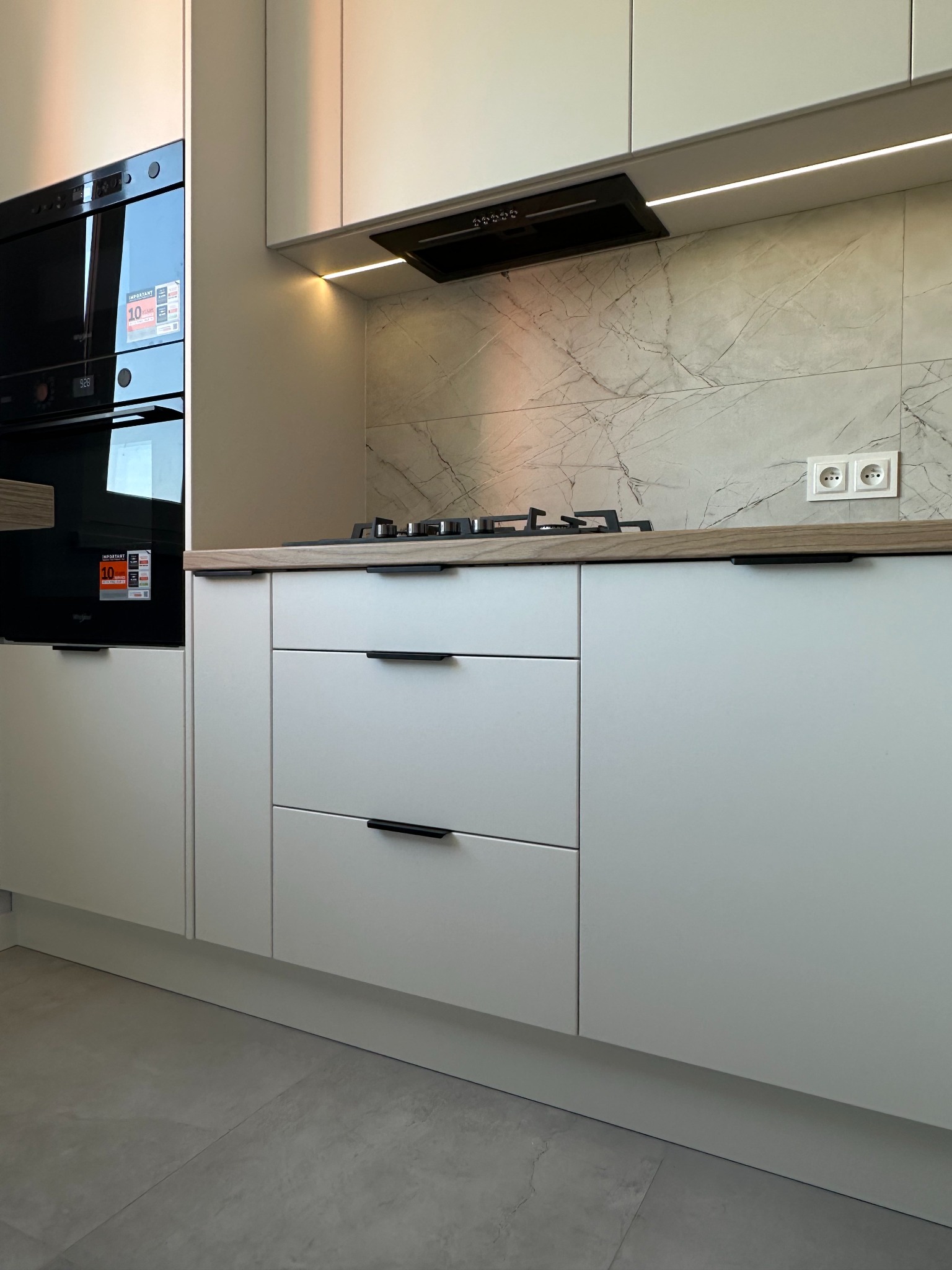 Nowoczesna kuchnia z białymi frontami, czarnymi uchwytami i marmurowym backsplash. Widoczne piekarniki w zabudowie i płyta gazowa. Oświetlenie LED podszafkowe.