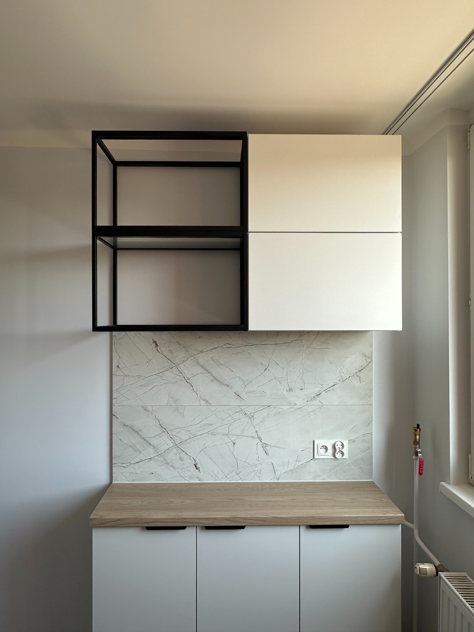 Nowoczesna zabudowa kuchenna: białe szafki dolne i górne z czarnym regałem. Blat drewnopodobny, ściana wykończona płytkami imitującymi marmur. Minimalistyczny design.