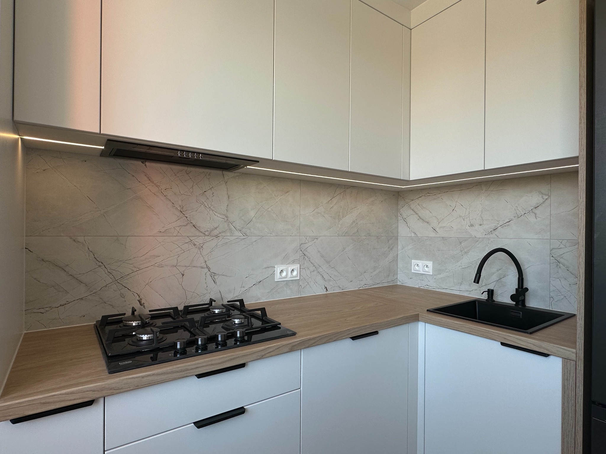 Biała, nowoczesna kuchnia z drewnianym blatem, marmurową ścianą i czarną armaturą. Uwagę przykuwa subtelne oświetlenie LED pod szafkami wiszącymi.