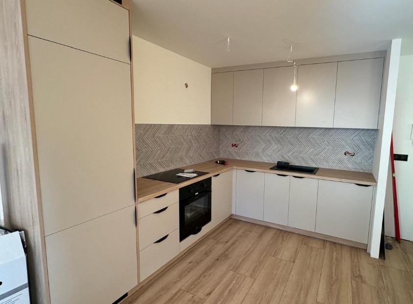 Jasna kuchnia z frontami w odcieniach beżu i szarości, drewniany blat i podłoga, wzorzysta ściana nad blatem. Widoczne płyty grzewcze i piekarnik.