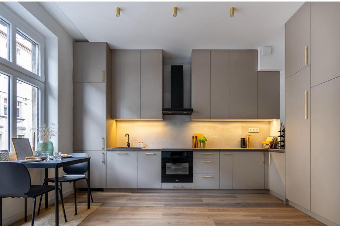 Nowoczesna kuchnia w odcieniach szarości z drewnianą podłogą, oświetleniem LED pod szafkami i złotymi uchwytami, stół z krzesłami przy oknie.