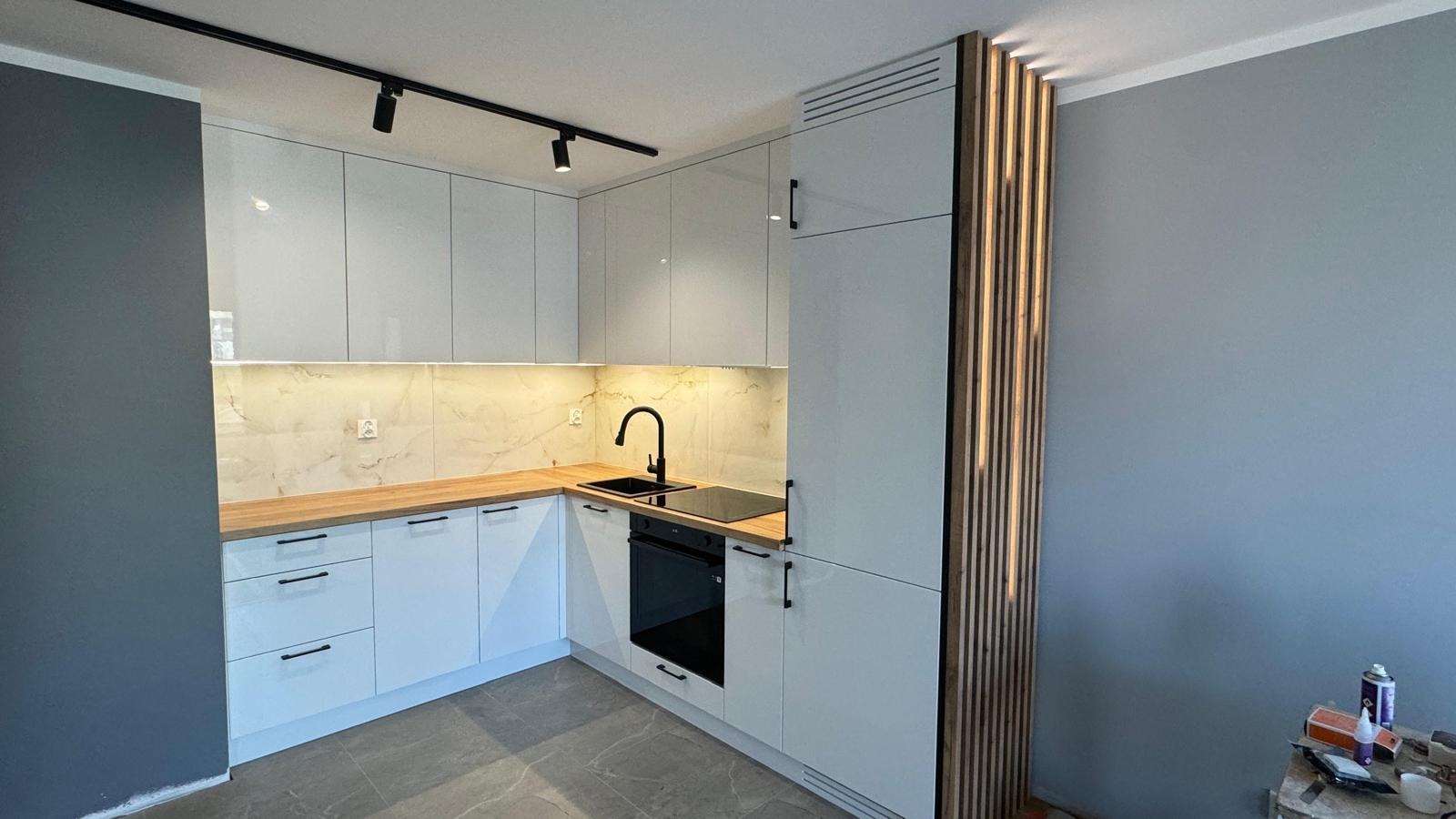 Nowoczesna kuchnia narożna z białymi, błyszczącymi frontami, drewnianym blatem i czarną armaturą. Uwagę zwraca pionowa, dekoracyjna ścianka z drewnianych listew.