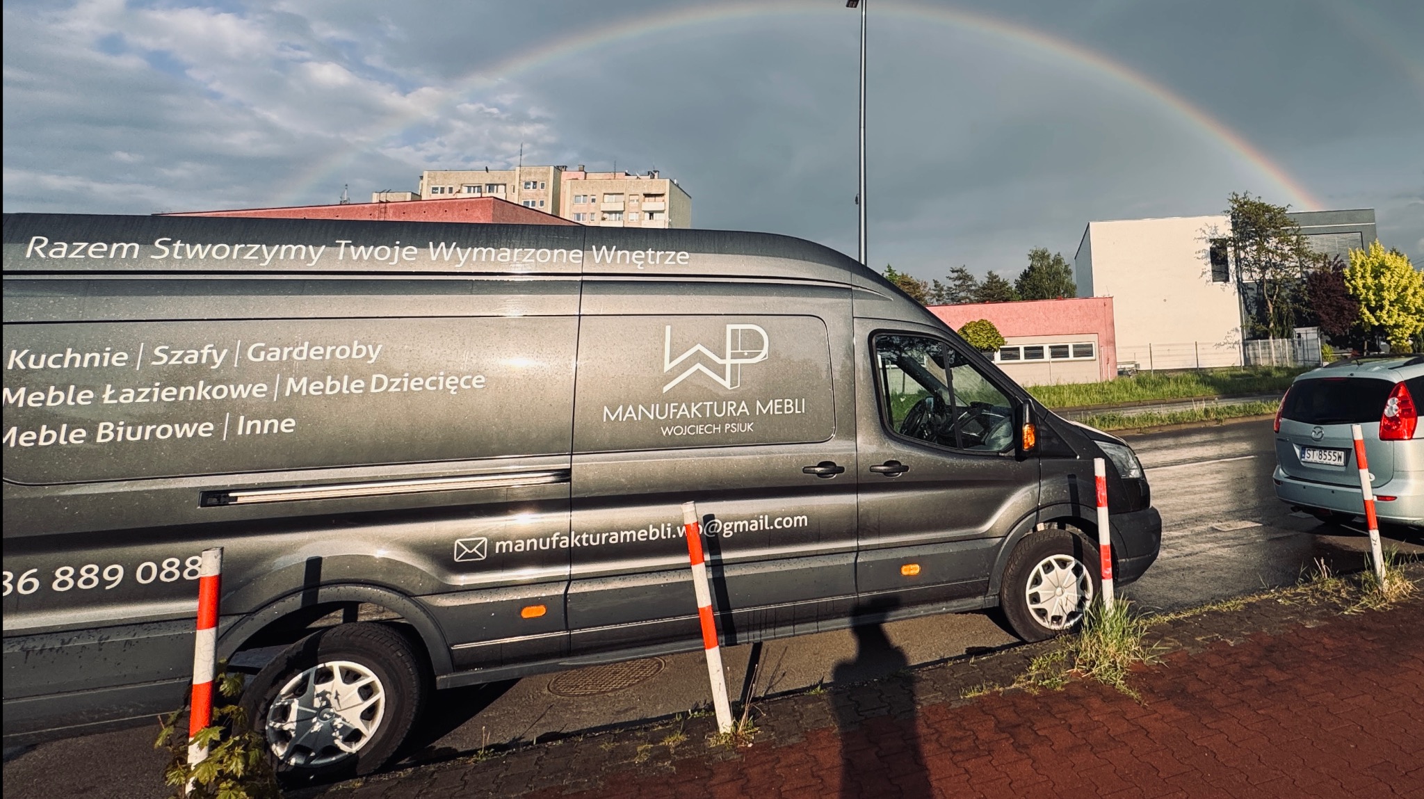 Szary bus firmy meblarskiej z tęczą w tle. Na pojeździe widoczne logo i informacje o ofercie: kuchnie, szafy, garderoby, meble łazienkowe, biurowe i dziecięce.