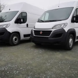 Dwa białe busy Fiat Ducato na żwirowym podłożu, ustawione blisko siebie, z widocznymi detalami przodu i boku jednego z pojazdów. Pochmurne niebo w tle.