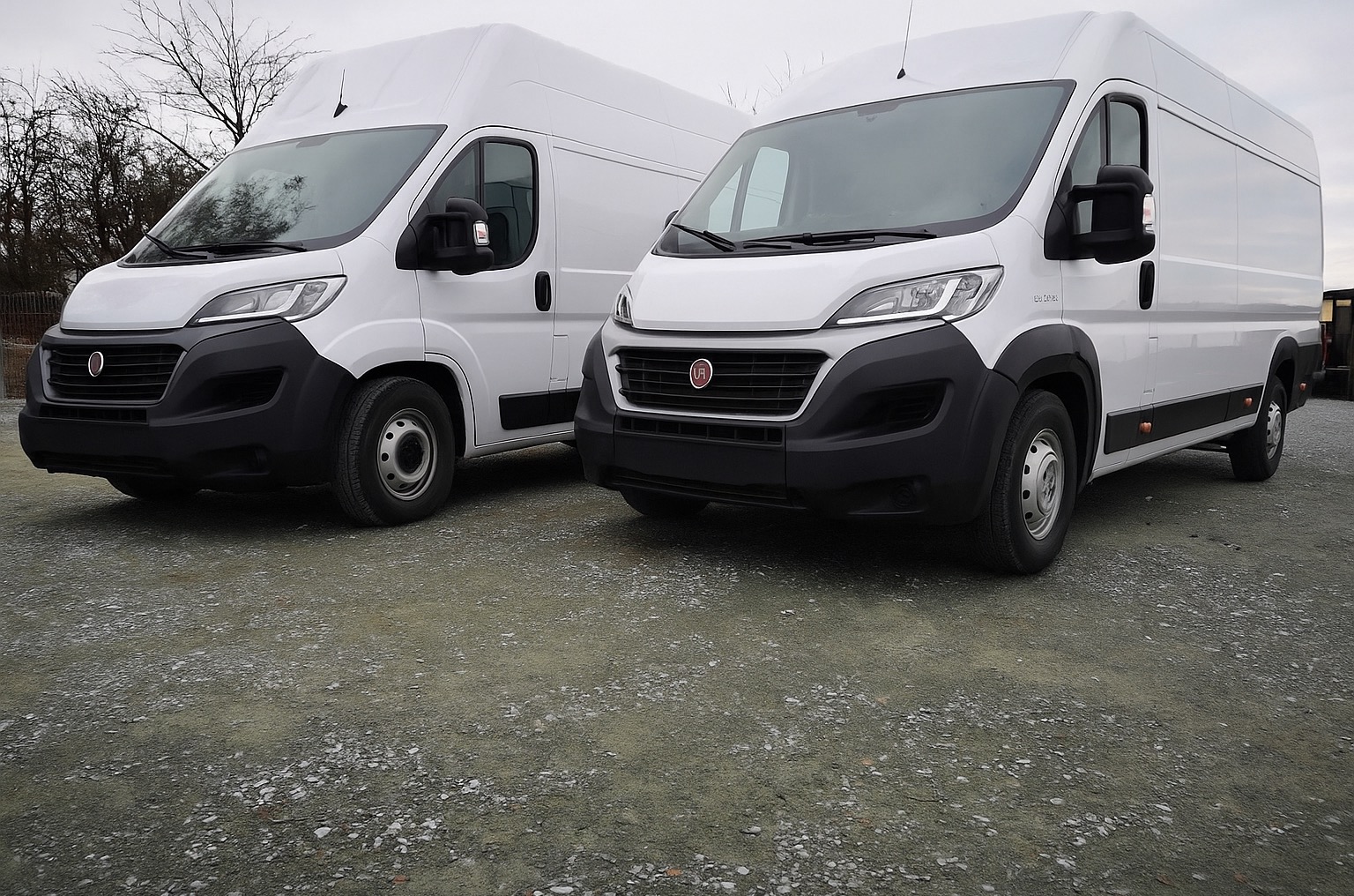 Dwa białe busy Fiat Ducato na żwirowym podłożu, ustawione blisko siebie, z widocznymi detalami przodu i boku jednego z pojazdów. Pochmurne niebo w tle.