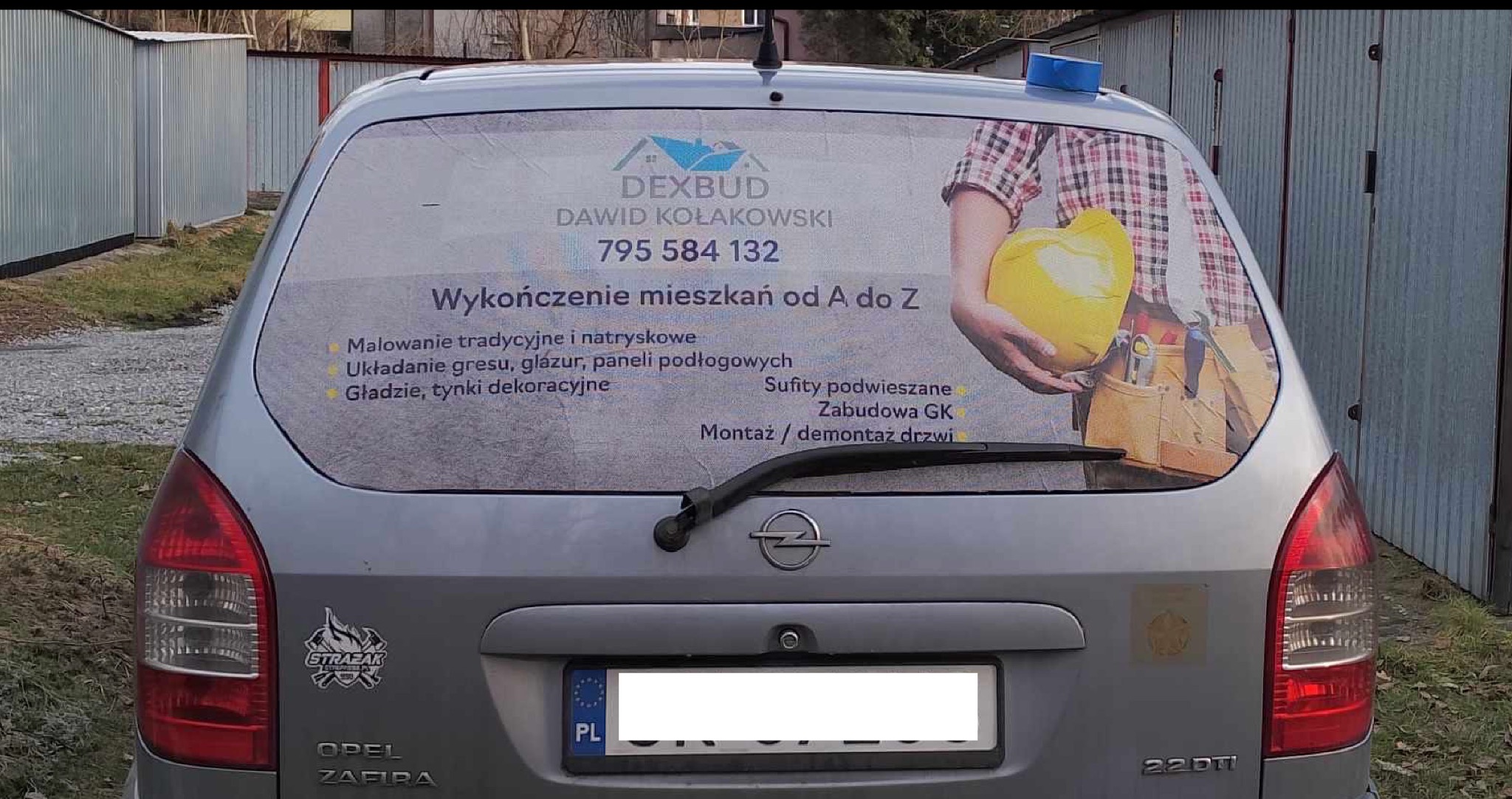Srebrny Opel Zafira z reklamą firmy Dexbud na tylnej szybie, oferującej wykończenie mieszkań od A do Z. Widoczne dane kontaktowe i zakres usług.