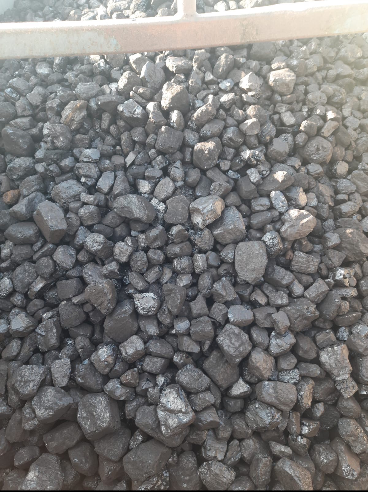 Kupa antracytu o nieregularnych kształtach, z widocznym metalowym elementem konstrukcyjnym w górnej części.  Światło podkreśla błyszczące powierzchnie bryłek.