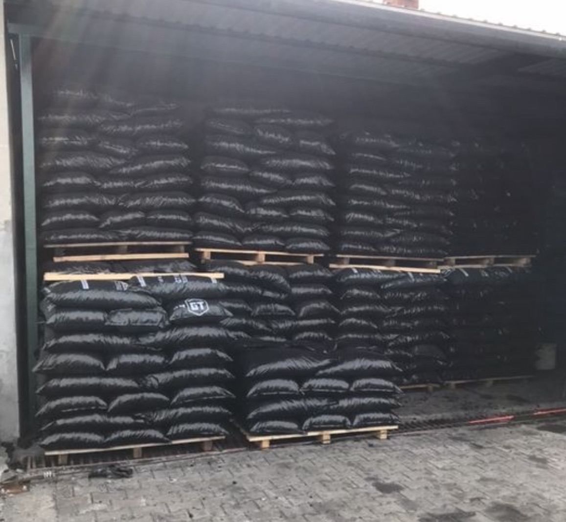 Magazyn z workami węgla ułożonymi na paletach pod zadaszeniem. Worki są czarne i szczelnie wypełnione. Widoczne logo producenta na workach.
