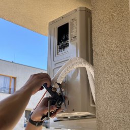 Coldmate Paulina Bochniak - Montaż zewnętrznej jednostki klimatyzacji na balkonie. Widoczne ręce instalatora przy pracy, rury w izolacji i fragment budynku w tle.