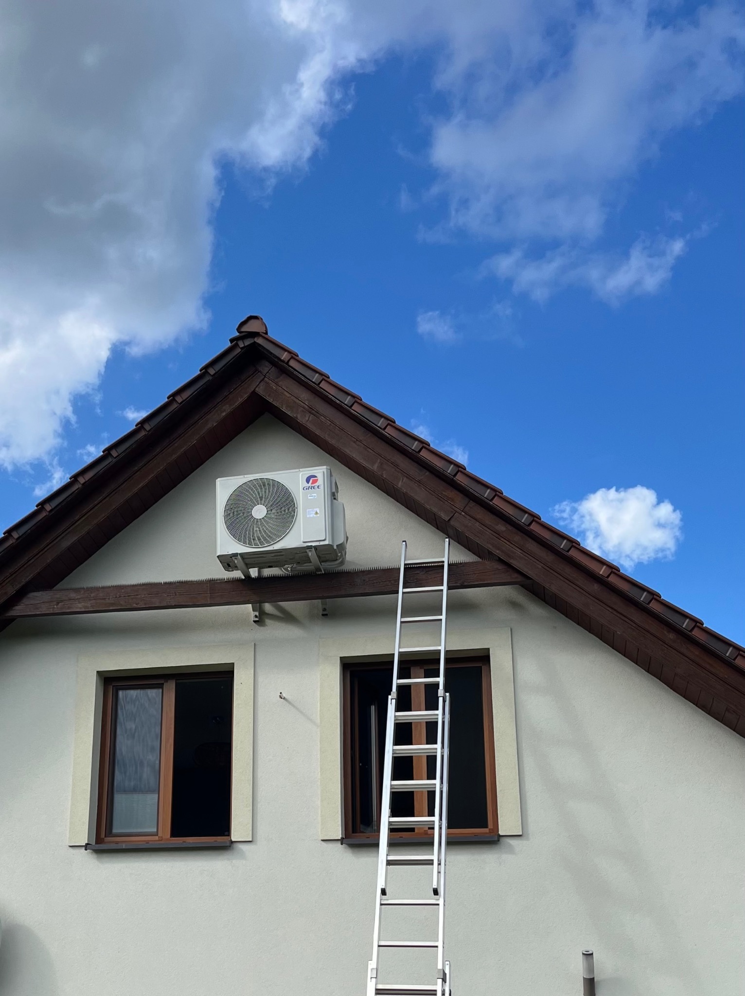 Biała jednostka zewnętrzna klimatyzacji Gree zamontowana na ścianie domu, obok okna. Przystawiona drabina sugeruje montaż lub serwis.