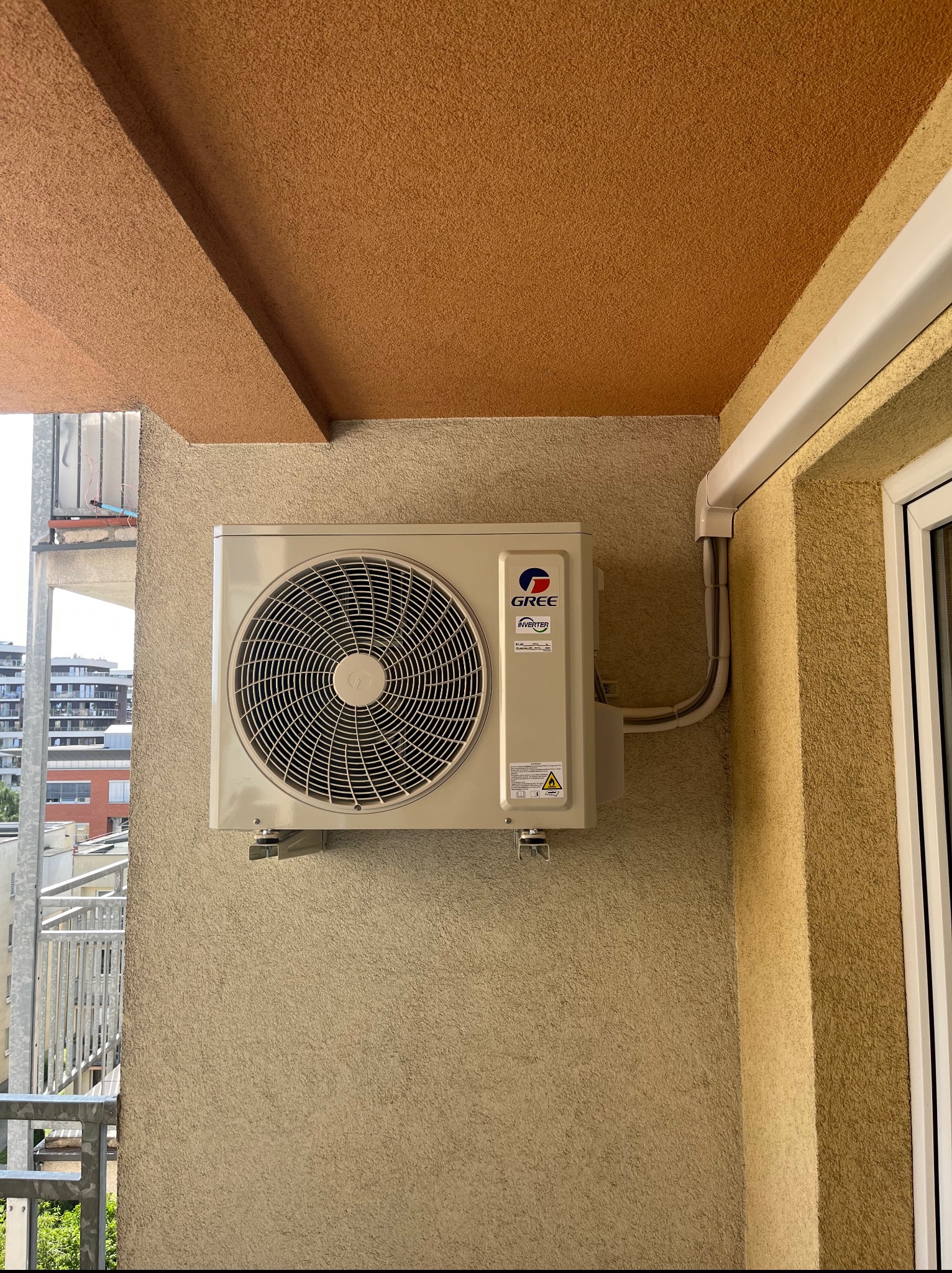 Zamontowana jednostka zewnętrzna klimatyzacji Gree na balkonie z widokiem na miasto, rury odprowadzające skropliny, beżowa elewacja budynku.