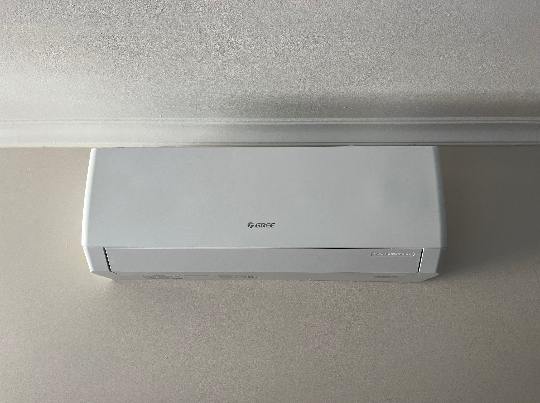 Biała klimatyzacja GREE zamontowana na ścianie pod sufitem z dekoracyjną listwą. Widok z przodu, z bliska, w neutralnym oświetleniu.