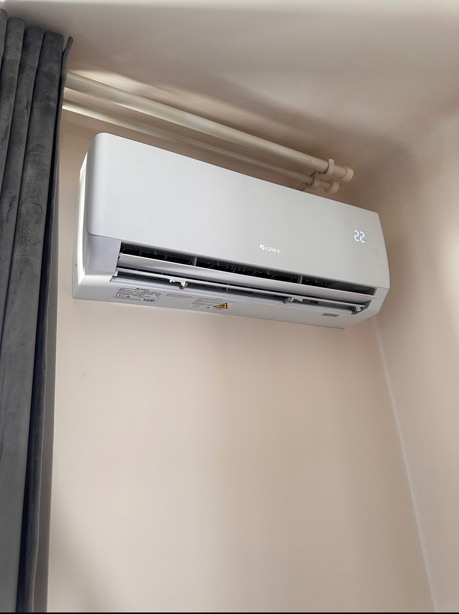 Biała klimatyzacja Gree na ścianie, ustawiona na 22 stopnie, widoczna część zasłony i rurki pod sufitem. Montaż klimatyzacji w domu.