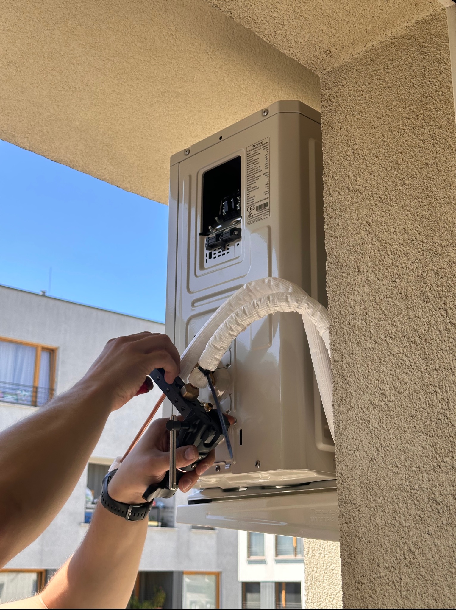Montaż zewnętrznej jednostki klimatyzacji na balkonie. Widoczne ręce instalatora przy pracy, rury w izolacji i fragment budynku w tle.