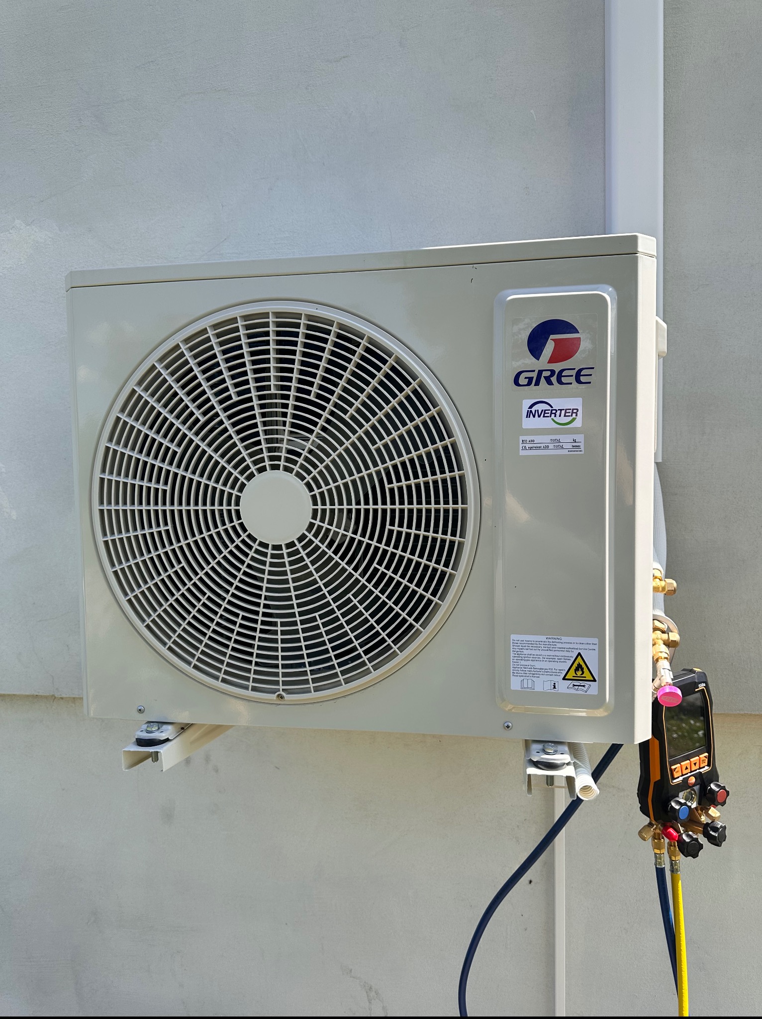Zewnętrzna jednostka klimatyzacji Gree zamontowana na ścianie, podłączona do narzędzi serwisowych. Widoczne przewody i manometr cyfrowy.