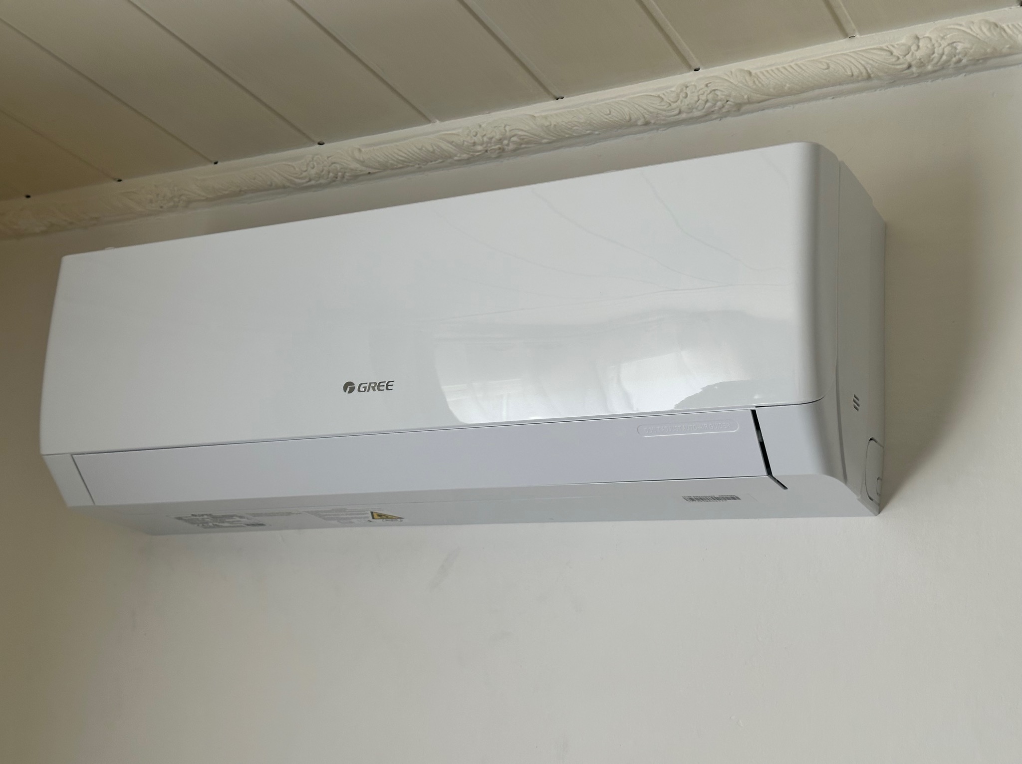Biała klimatyzacja marki Gree zamontowana na kremowej ścianie, widoczny ozdobny gzyms sufitowy. Ujęcie z dołu, lekko z prawej strony.