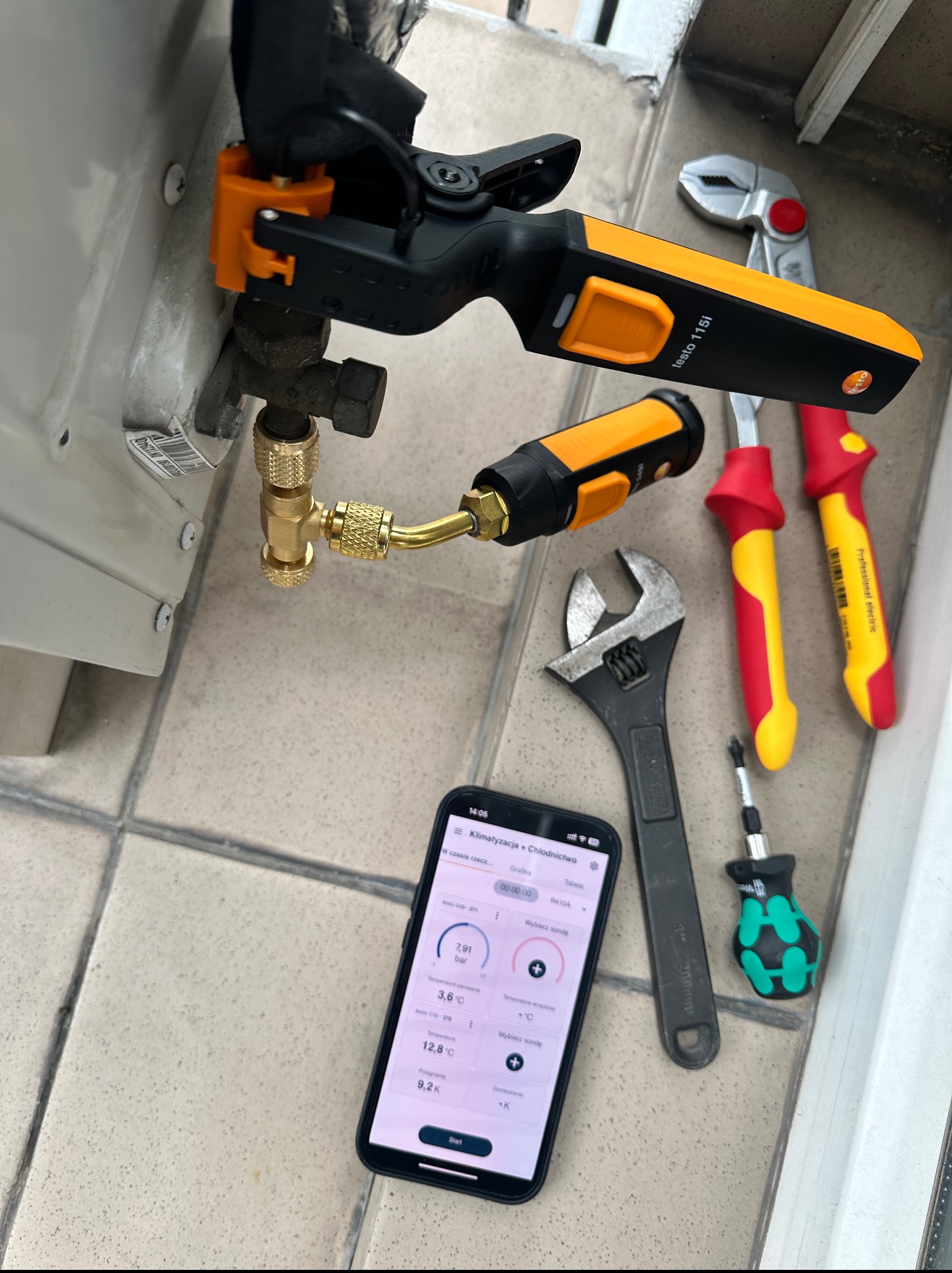 Pomiar parametrów klimatyzacji narzędziem Testo 115i, smartfon z aplikacją, klucze, kombinerki na tle płytek. Diagnostyka klimatyzacji w trakcie serwisu.