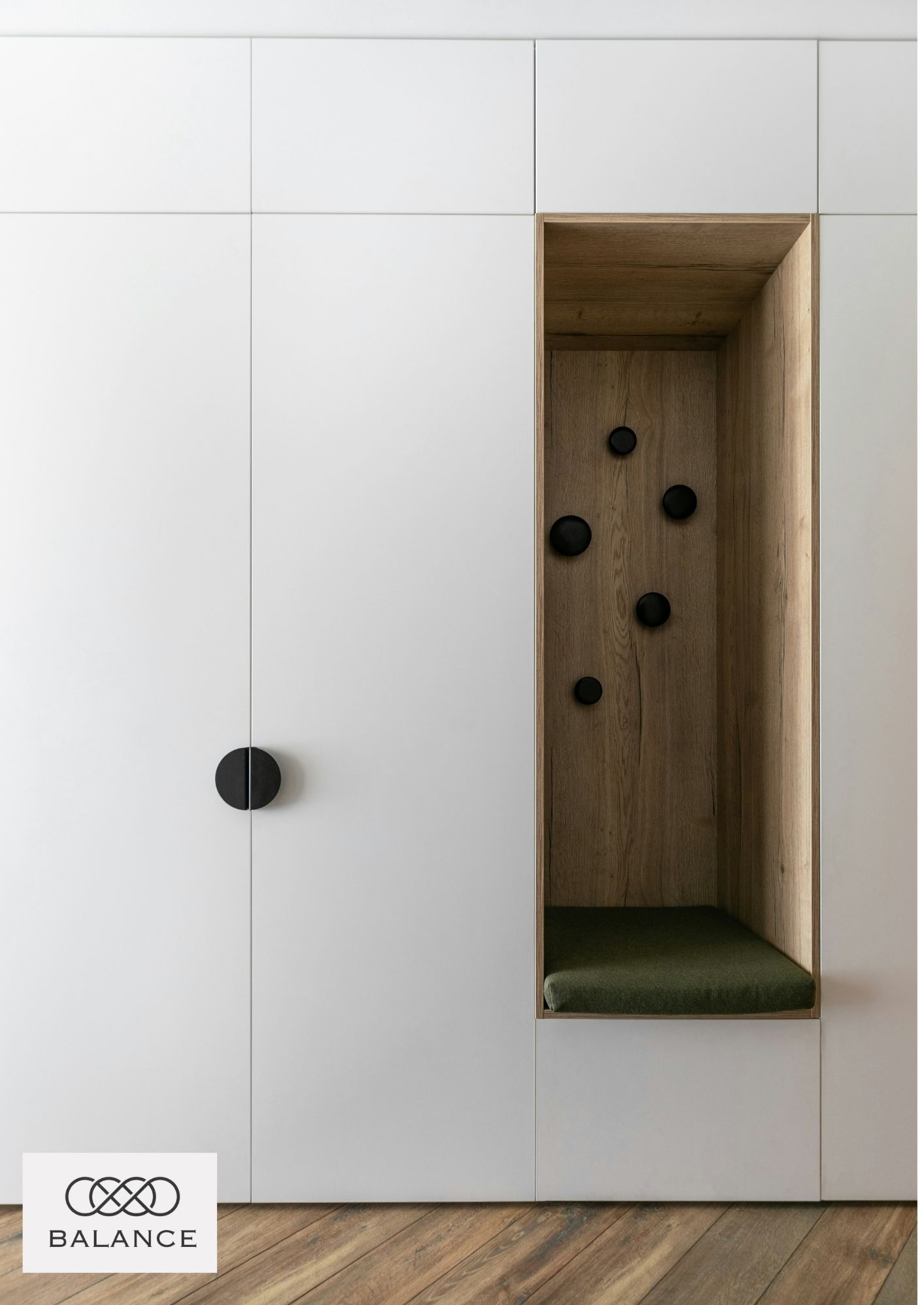 Biała szafa wnękowa z drewnianą wnęką z siedziskiem i czarnymi haczykami. Minimalistyczny design, jasne wnętrze, podłoga z paneli.