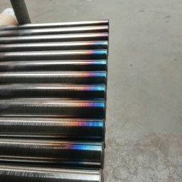 TOMASZ KULIGOWSKI DAR-TOM - Rząd metalowych prętów z opalizującym zabarwieniem na końcach, leżących na szarej powierzchni. Widoczne ślady obróbki.