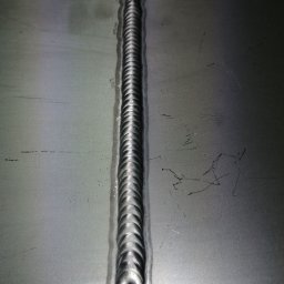 TOMASZ KULIGOWSKI DAR-TOM - Detal spoiny na metalowej powierzchni. Widoczna regularna struktura spawu, z drobnymi niedoskonałościami i zarysowaniami na materiale. Metal ma szary, surowy odcień.