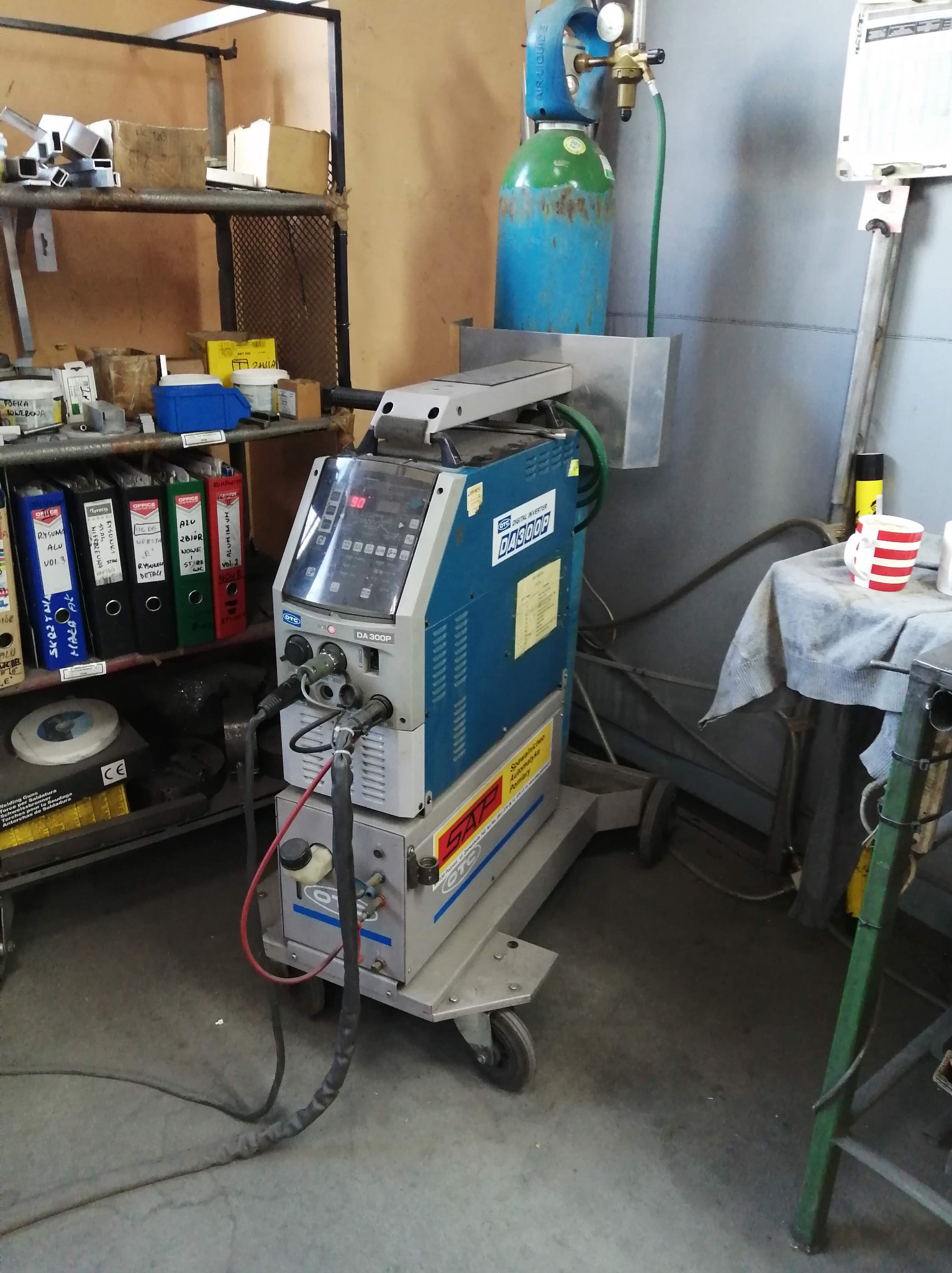 Mobilna spawarka inwertorowa DA300P z butlą gazową w warsztacie, na tle półek z dokumentami i narzędziami. Widoczny kabel i elementy sterujące urządzenia.