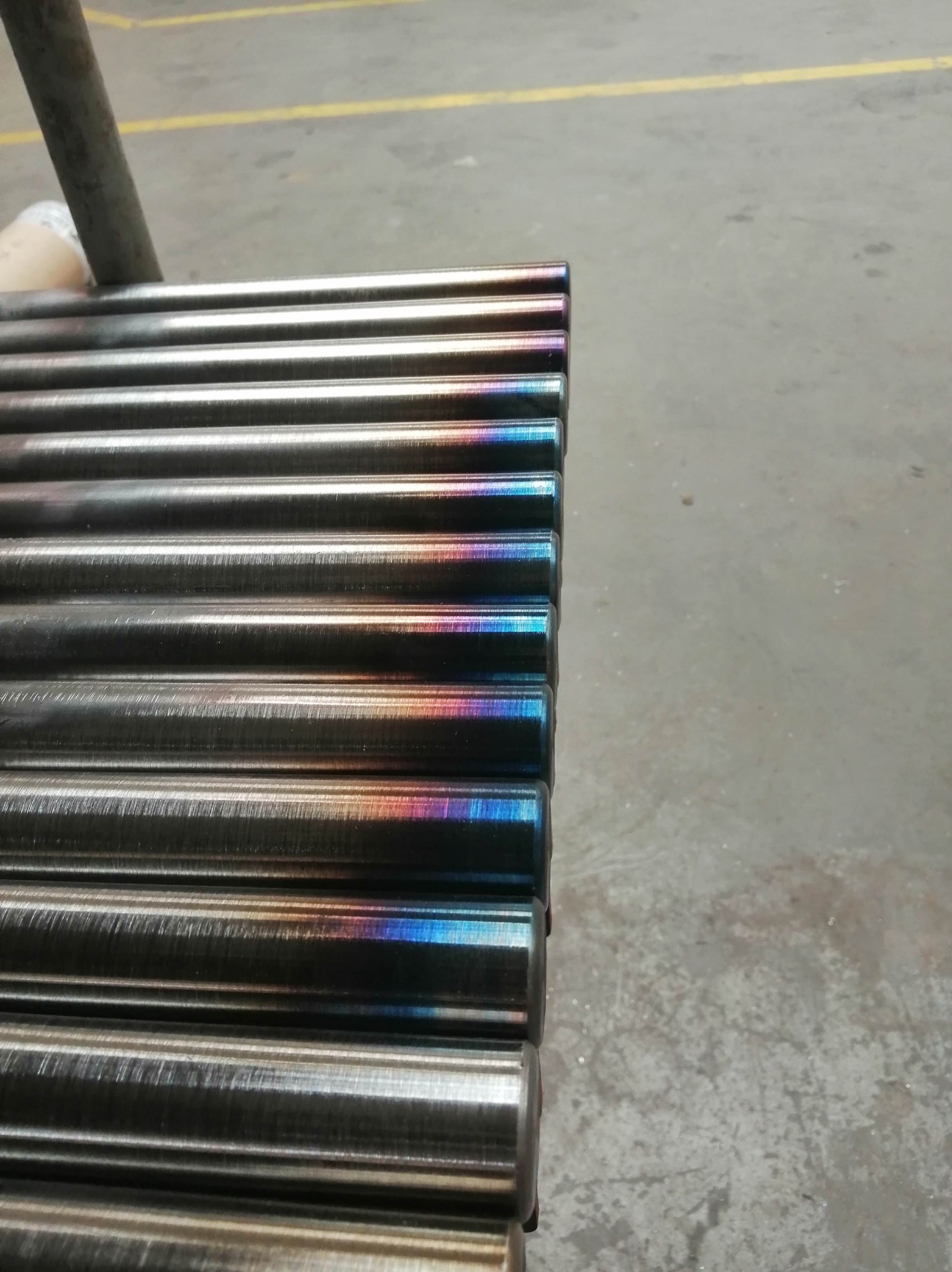 Rząd metalowych prętów z opalizującym zabarwieniem na końcach, leżących na szarej powierzchni. Widoczne ślady obróbki.