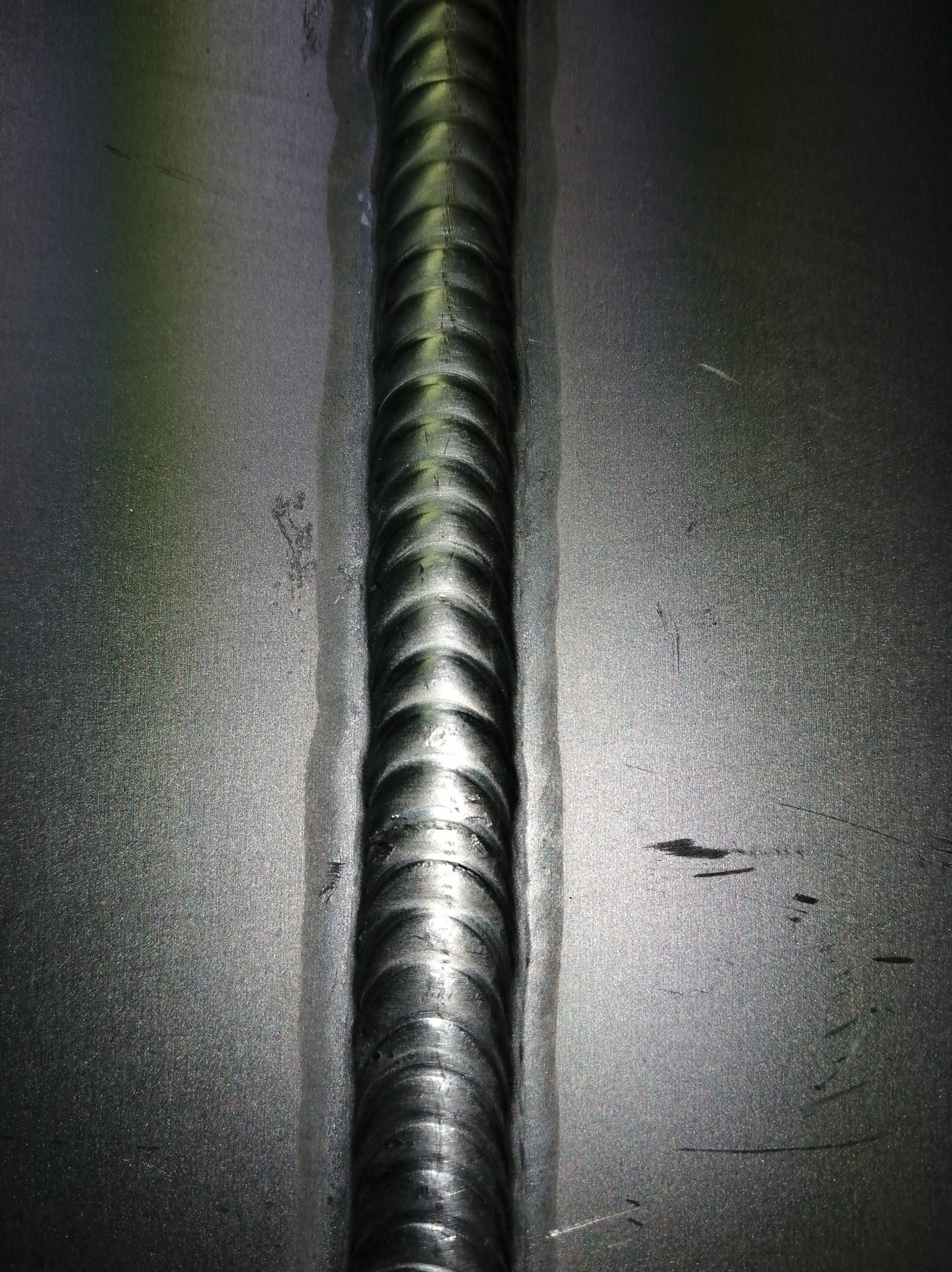 Precyzyjne, pionowe złącze spawane na metalowej powierzchni, ukazujące regularne, łuskowate ślady spawu i delikatne przebarwienia termiczne wokół spoiny.