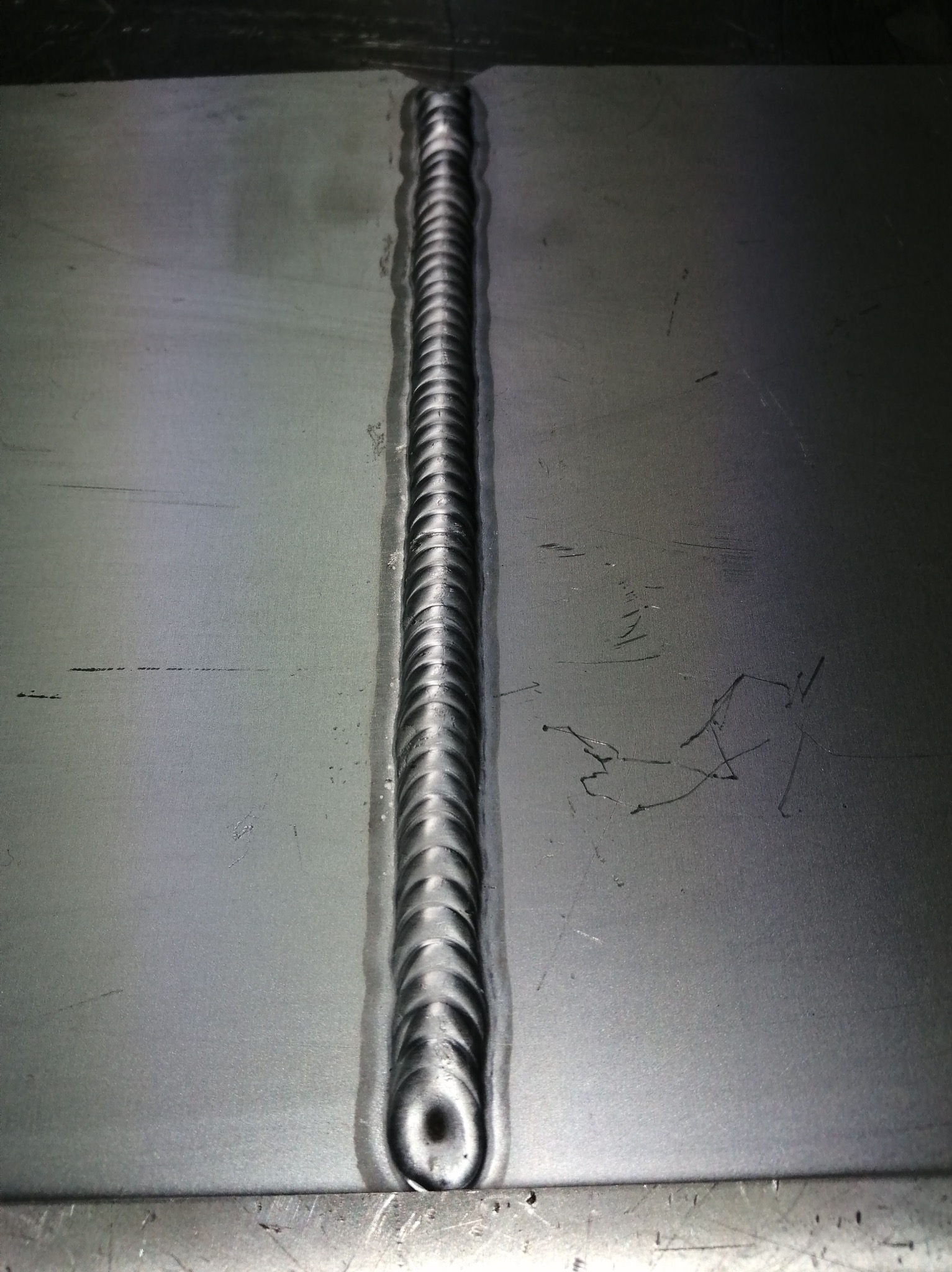 Detal spoiny na metalowej powierzchni. Widoczna regularna struktura spawu, z drobnymi niedoskonałościami i zarysowaniami na materiale. Metal ma szary, surowy odcień.