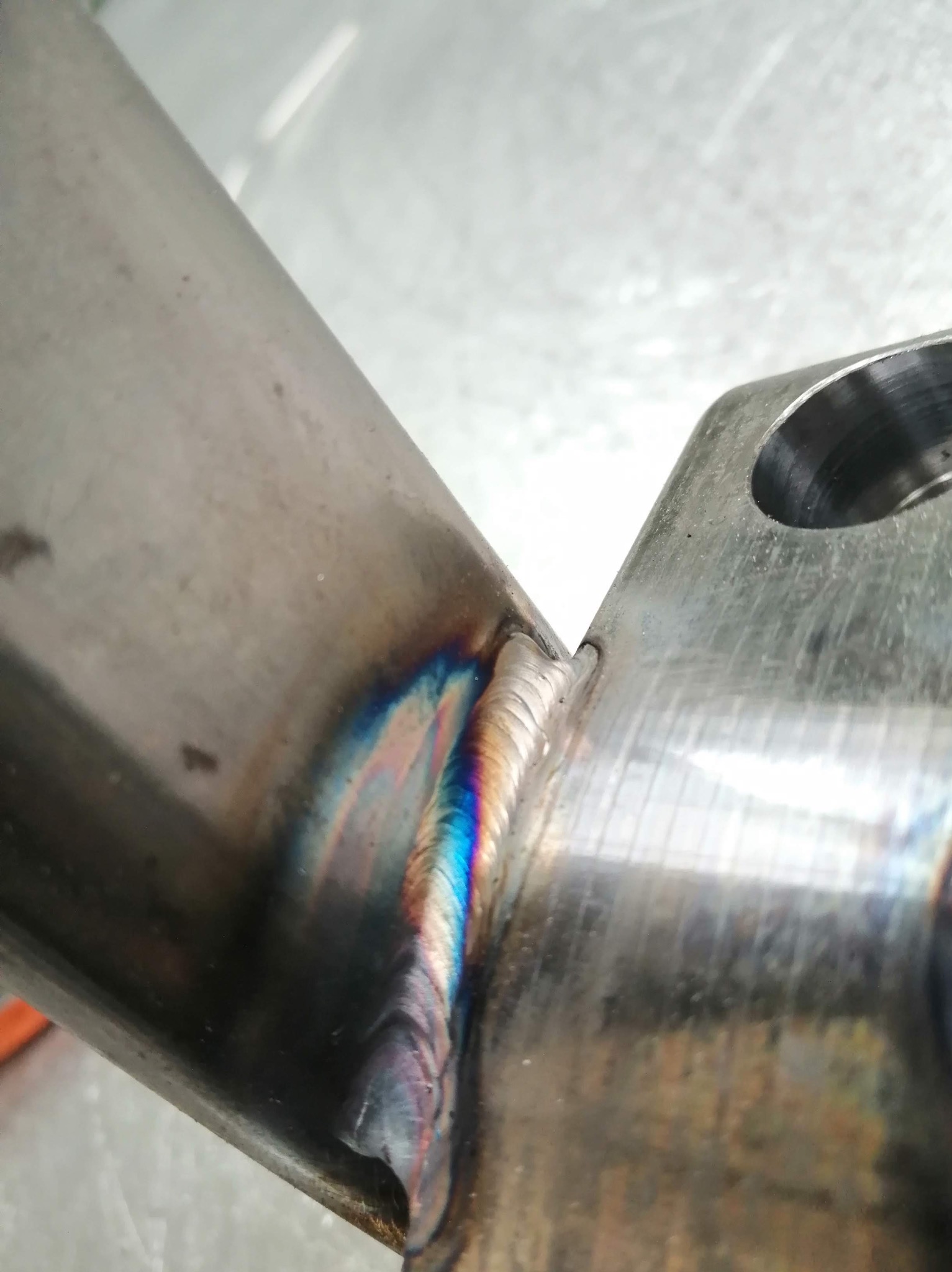 Spoina łącząca metalowy profil z płaskownikiem, widoczne tęczowe zabarwienia po spawaniu. Detal konstrukcji spawanej z bliska, z otworem na śrubę.