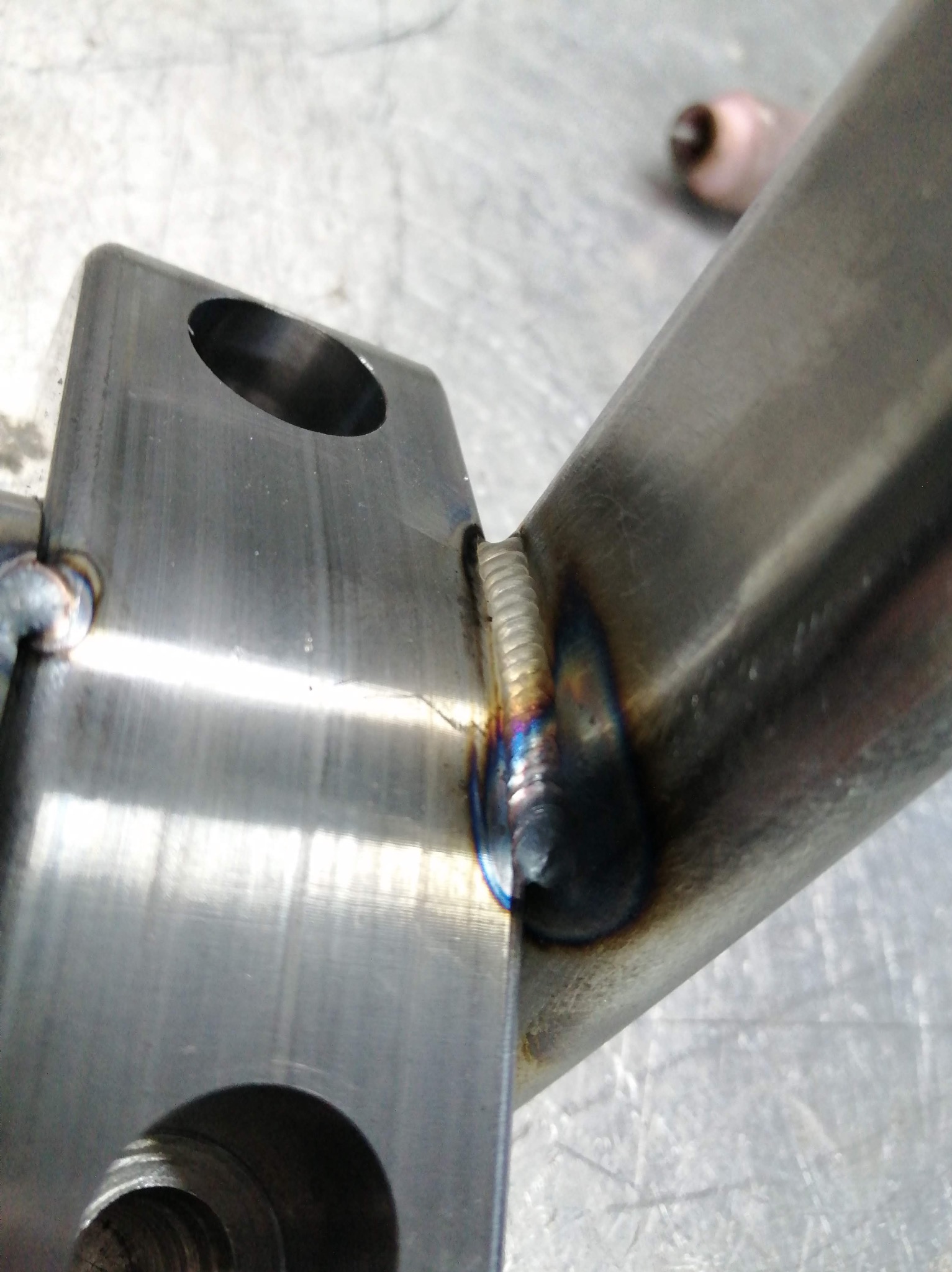 Spoina łącząca dwa elementy metalowe, widoczne przebarwienia termiczne na spoinie, co wskazuje na świeżość i jakość wykonania, na szlifowanym tle.