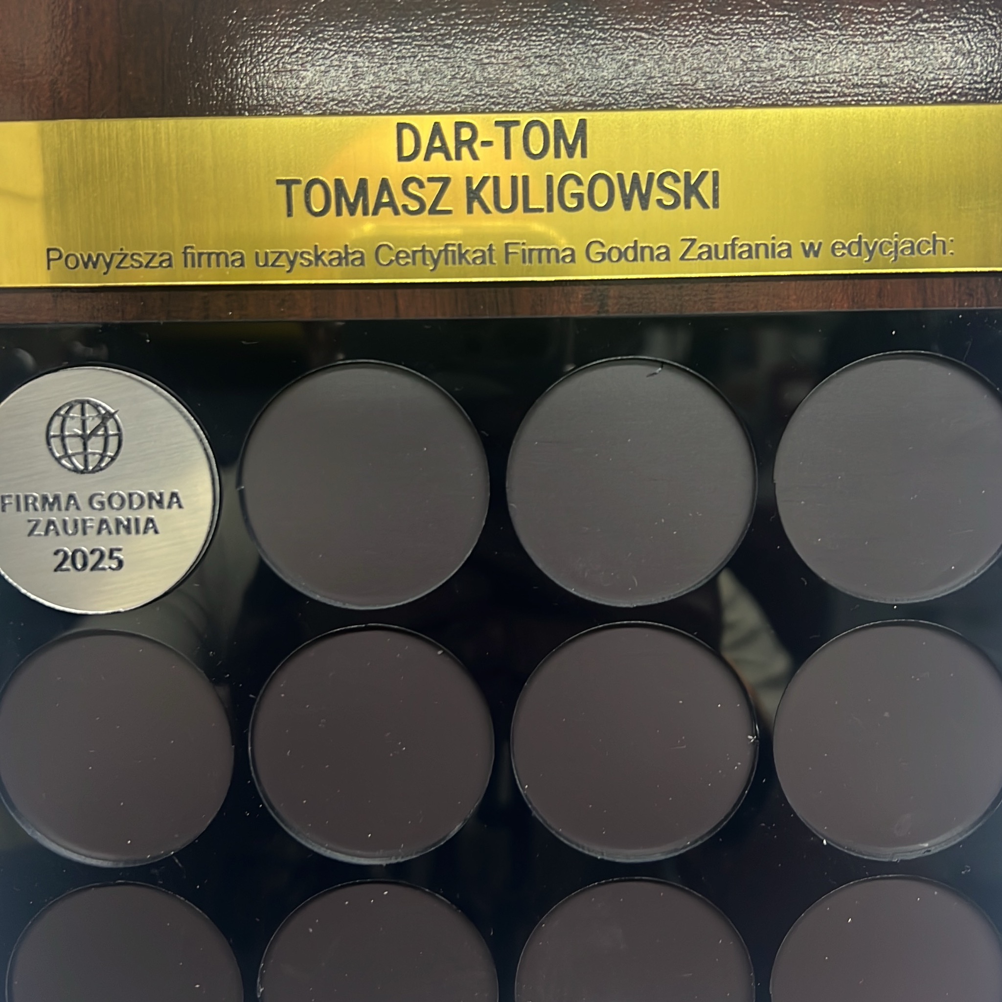 Tablica z certyfikatem 'Firma Godna Zaufania' dla DAR-TOM Tomasz Kuligowski, edycja 2025. Metalowa plakietka z logo i nazwą firmy. Widoczne puste miejsca na kolejne certyfikaty.