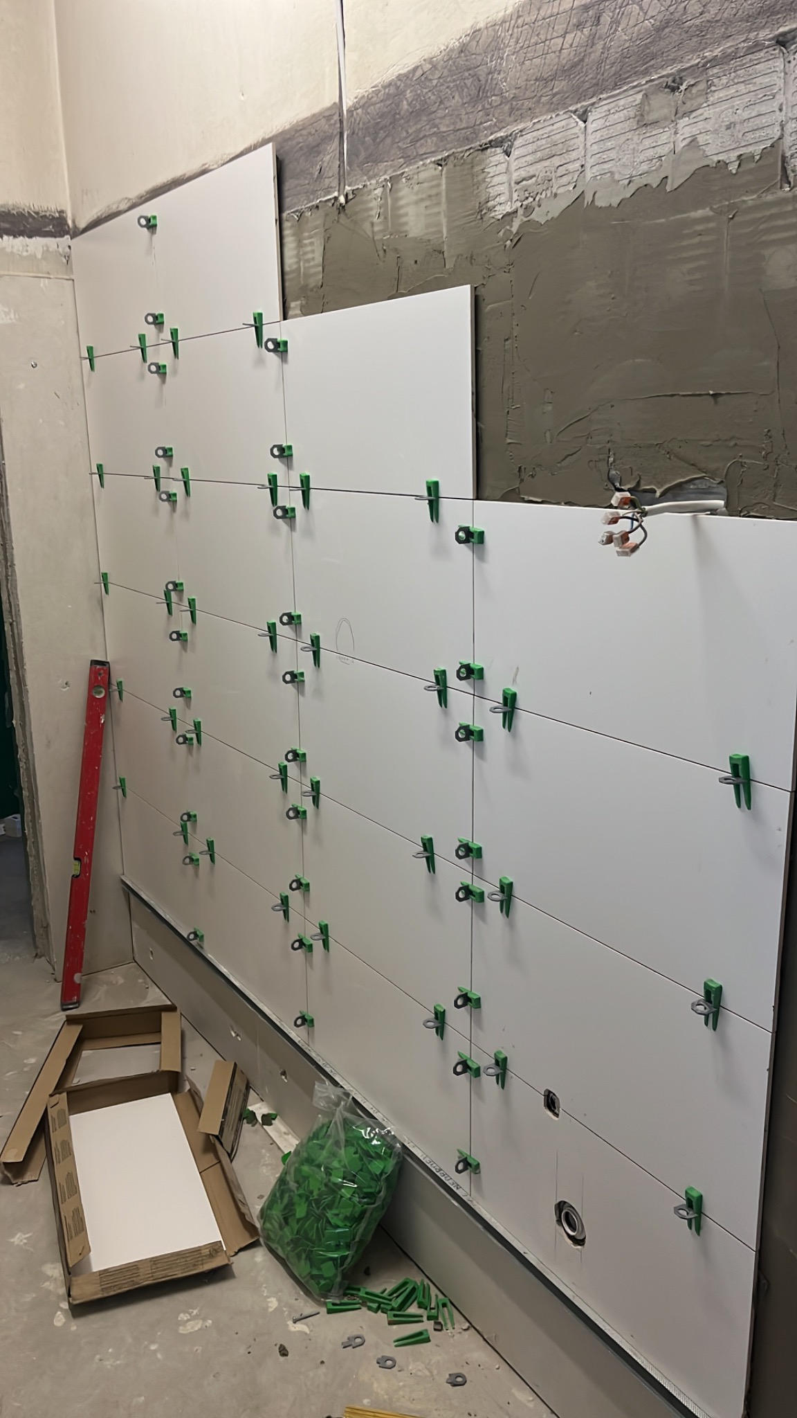 Białe płytki na ścianie, mocowane zielonymi klipsami. Częściowo przyklejone, widoczny klej. Poziomica i karton z płytkami na podłodze.