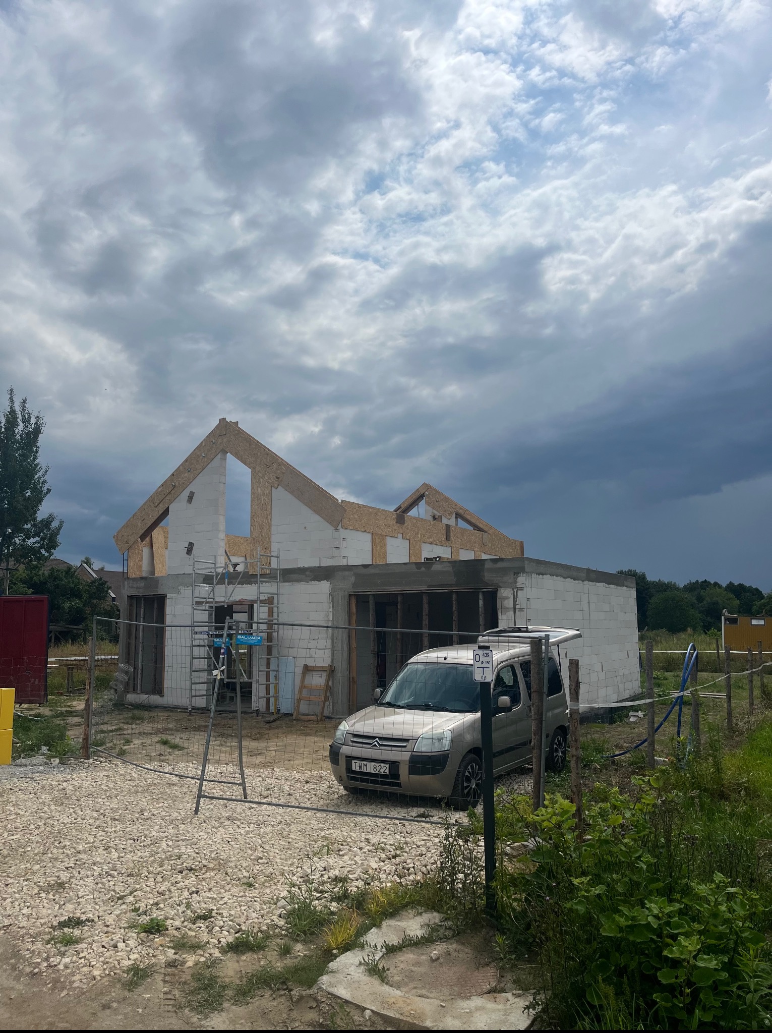 Budowa domu jednorodzinnego w stanie surowym otwartym. Ściany z betonu komórkowego, więźba dachowa, auto osobowe na posesji, pochmurne niebo.