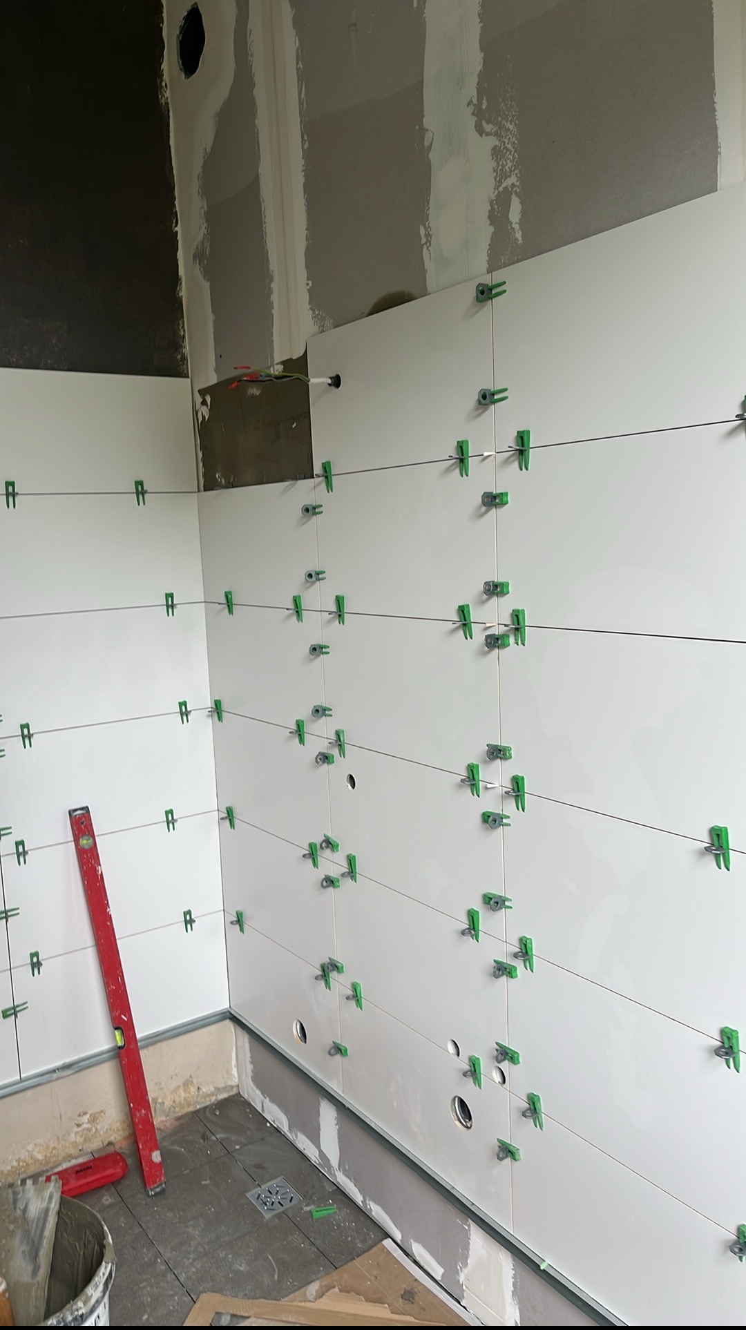 Białe płytki ścienne w trakcie montażu w łazience, z użyciem klipsów poziomujących i poziomicy. Widoczne przygotowane podłoże i otwory na instalacje.