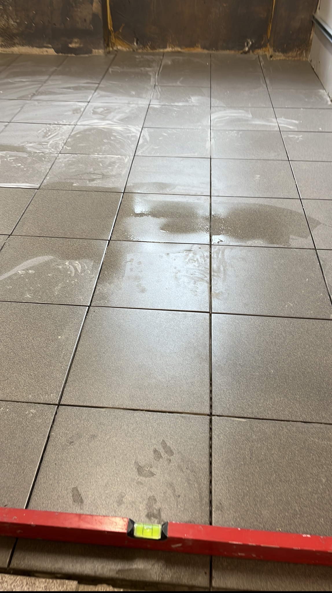 Ułożone, szare płytki podłogowe w trakcie prac. Widoczna poziomica i ślady po fugowaniu. Tło stanowi surowa ściana w trakcie wykończenia.