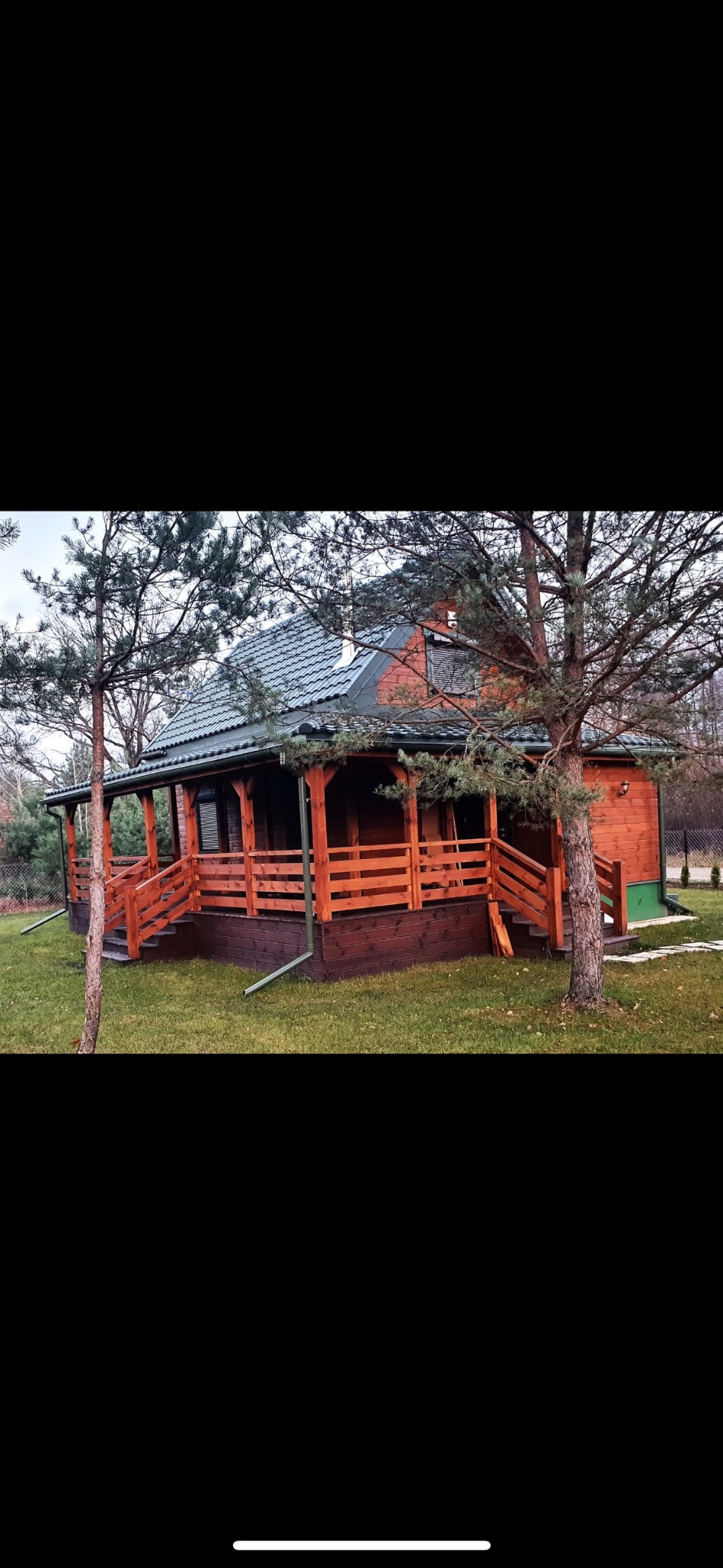 Drewniany dom z werandą i szarym dachem na trawniku, otoczony drzewami. Widoczne rynny i schody wejściowe. Całość w naturalnej kolorystyce.