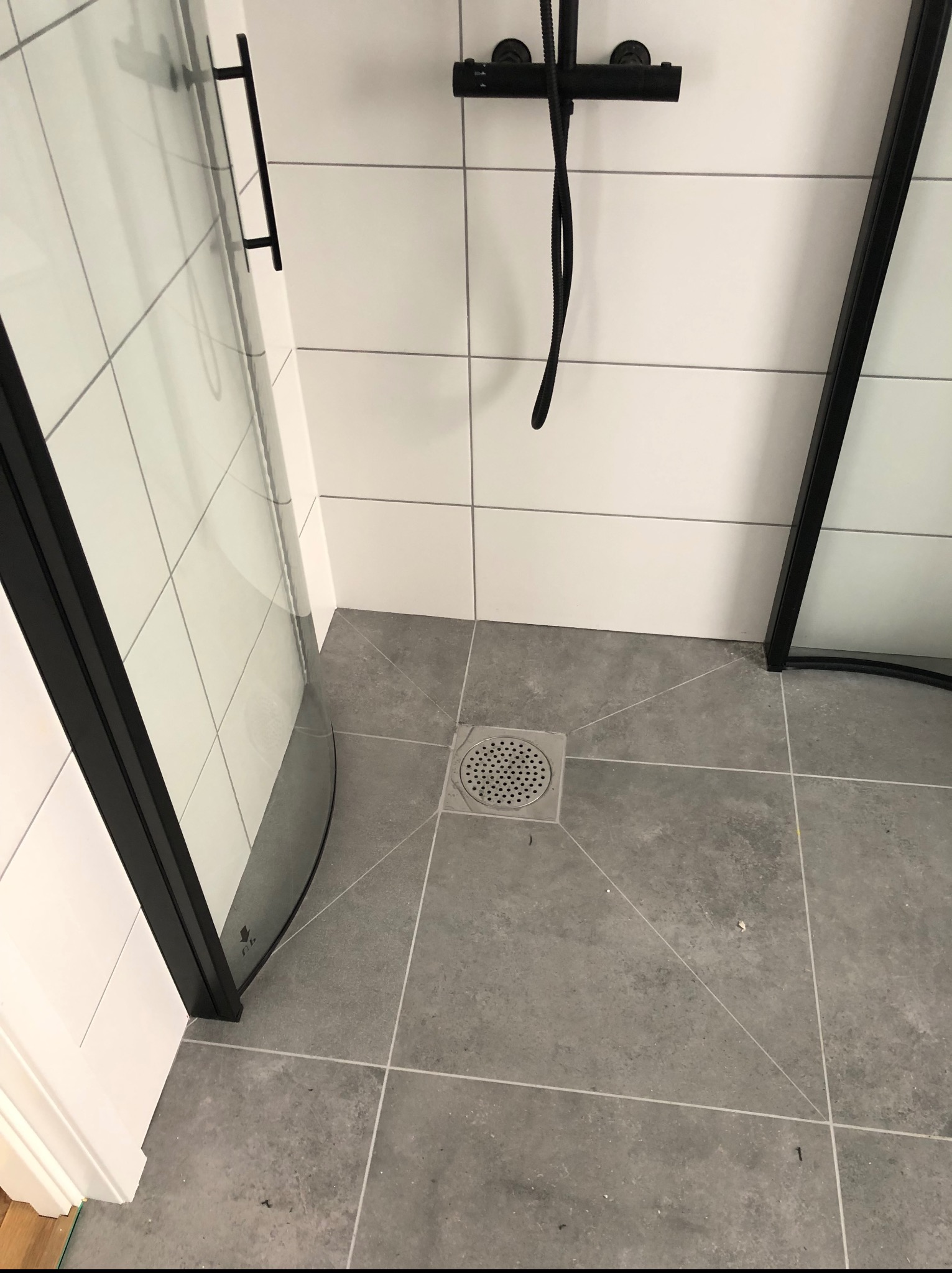 Szara, kwadratowa płytka podłogowa w nowoczesnej łazience z czarną armaturą i kabiną prysznicową z czarnymi ramami. Widoczny odpływ liniowy.
