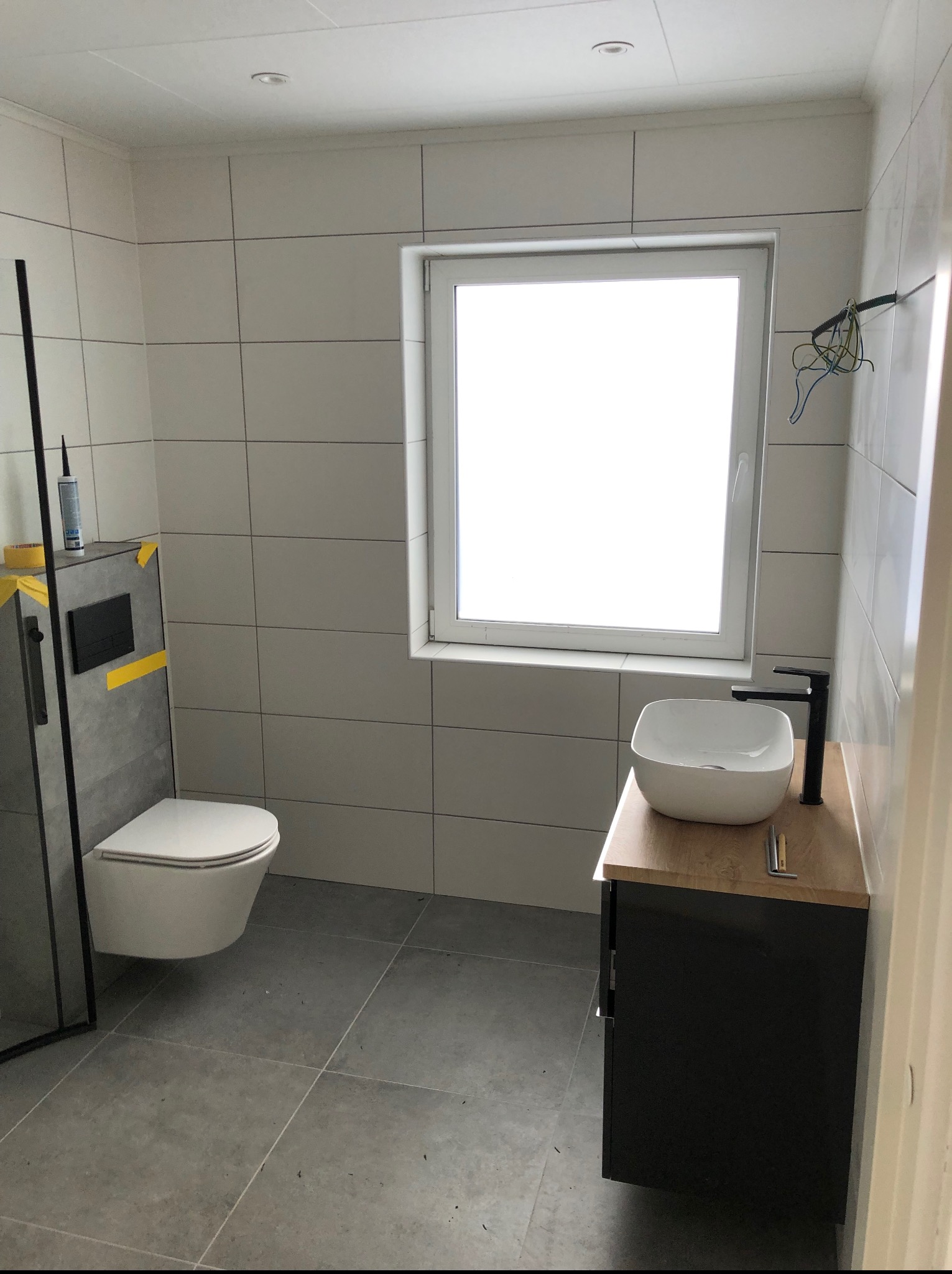 Nowoczesna łazienka z białymi płytkami, podwieszaną toaletą, umywalką na drewnianym blacie i czarną armaturą. Widoczne narzędzia i silikon. Minimalistyczny design.