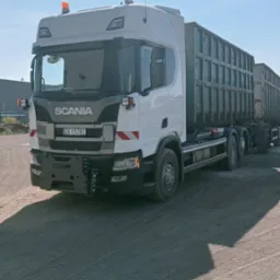 Biała ciężarówka Scania z dwoma kontenerami na gruz, zaparkowana na piaszczystym terenie w słoneczny dzień. Widoczne pomarańczowe światła ostrzegawcze.