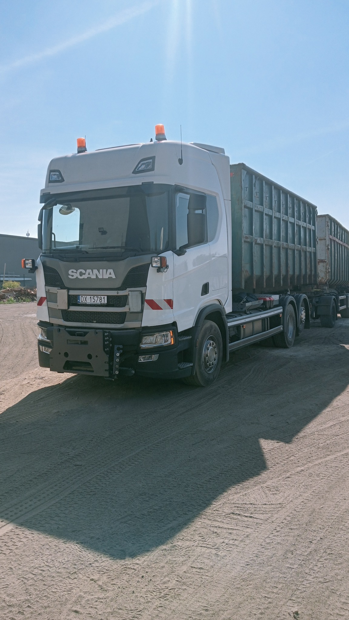 Biała ciężarówka Scania z dwoma kontenerami na gruz, zaparkowana na piaszczystym terenie w słoneczny dzień. Widoczne pomarańczowe światła ostrzegawcze.