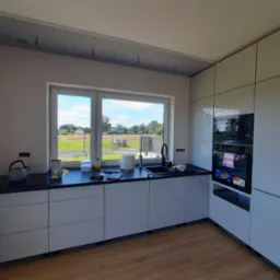 Jasna kuchnia z białymi frontami, czarnym blatem i widokiem na zielone pole z okna. Nowoczesny design z piekarnikiem w zabudowie i drewnianą podłogą.