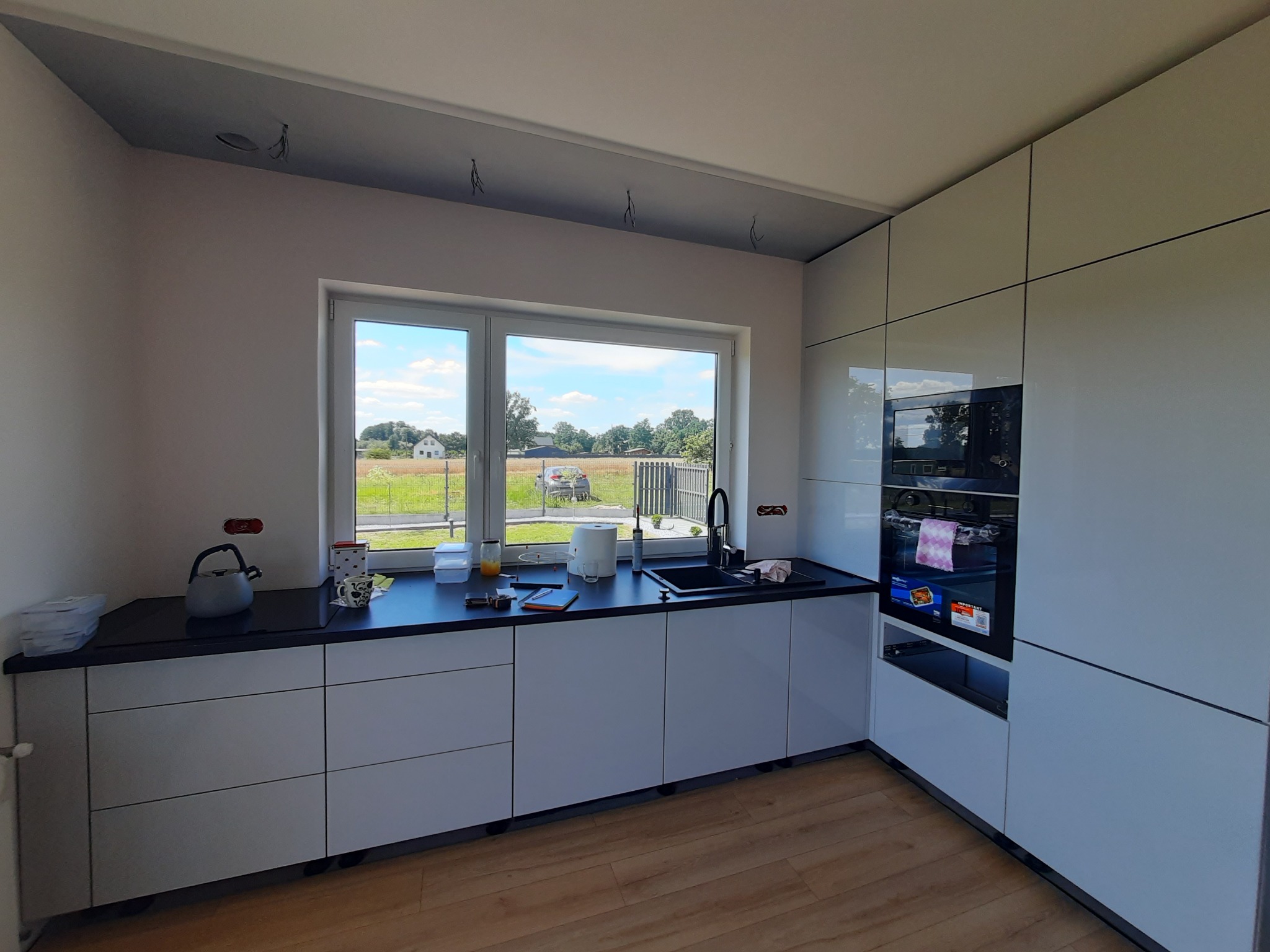 Jasna kuchnia z białymi frontami, czarnym blatem i widokiem na zielone pole z okna. Nowoczesny design z piekarnikiem w zabudowie i drewnianą podłogą.