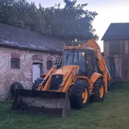 Żółta koparko-ładowarka JCB na trawie przed budynkiem z cegły i drewnianą stodołą w tle, uchwycona o zmierzchu.